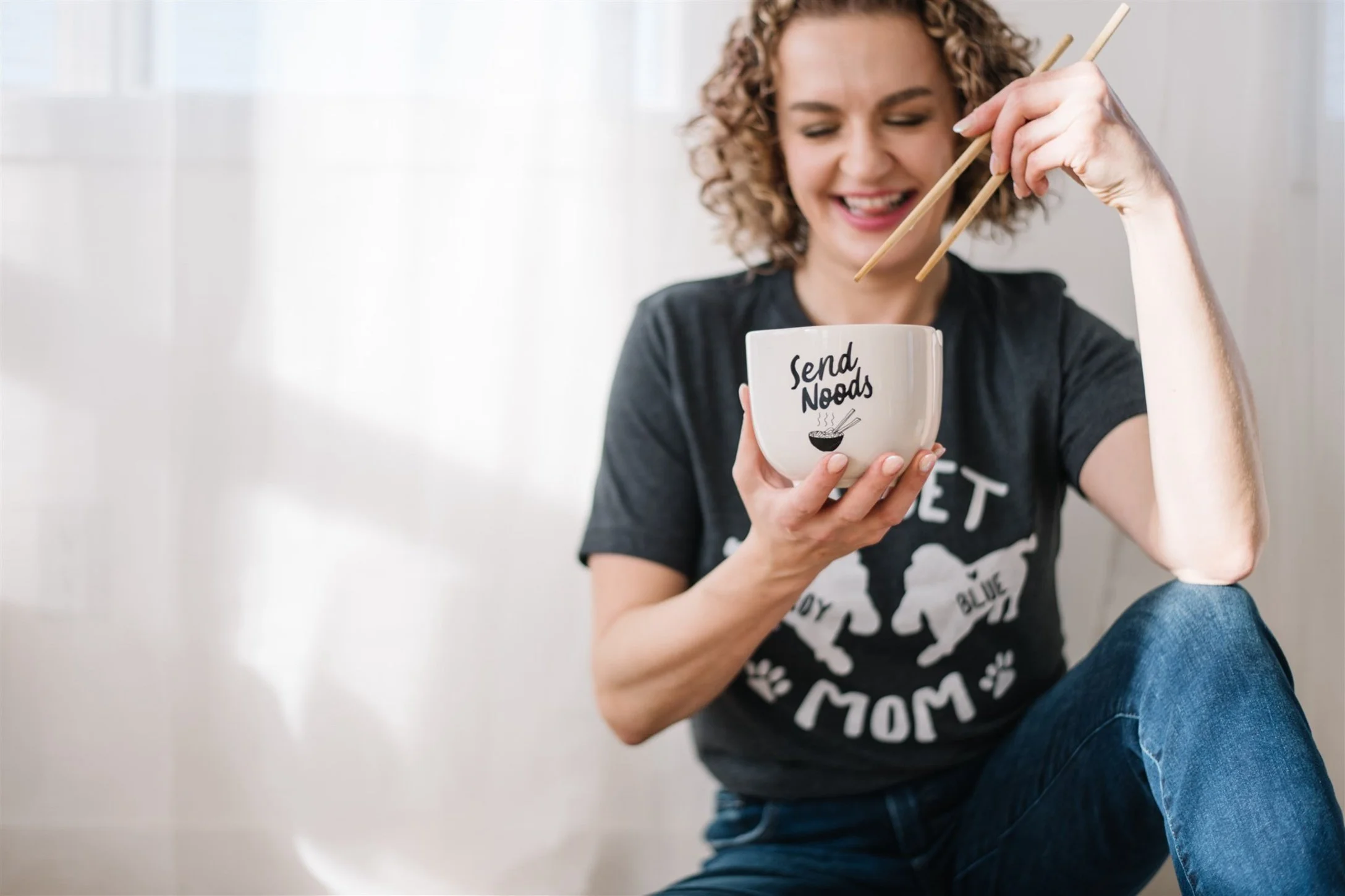 Maggie Nowak, owner of Story Within in Edmonton sitting on the floor, smiling, holding a bowl labeled "Send Noods," using chopsticks.