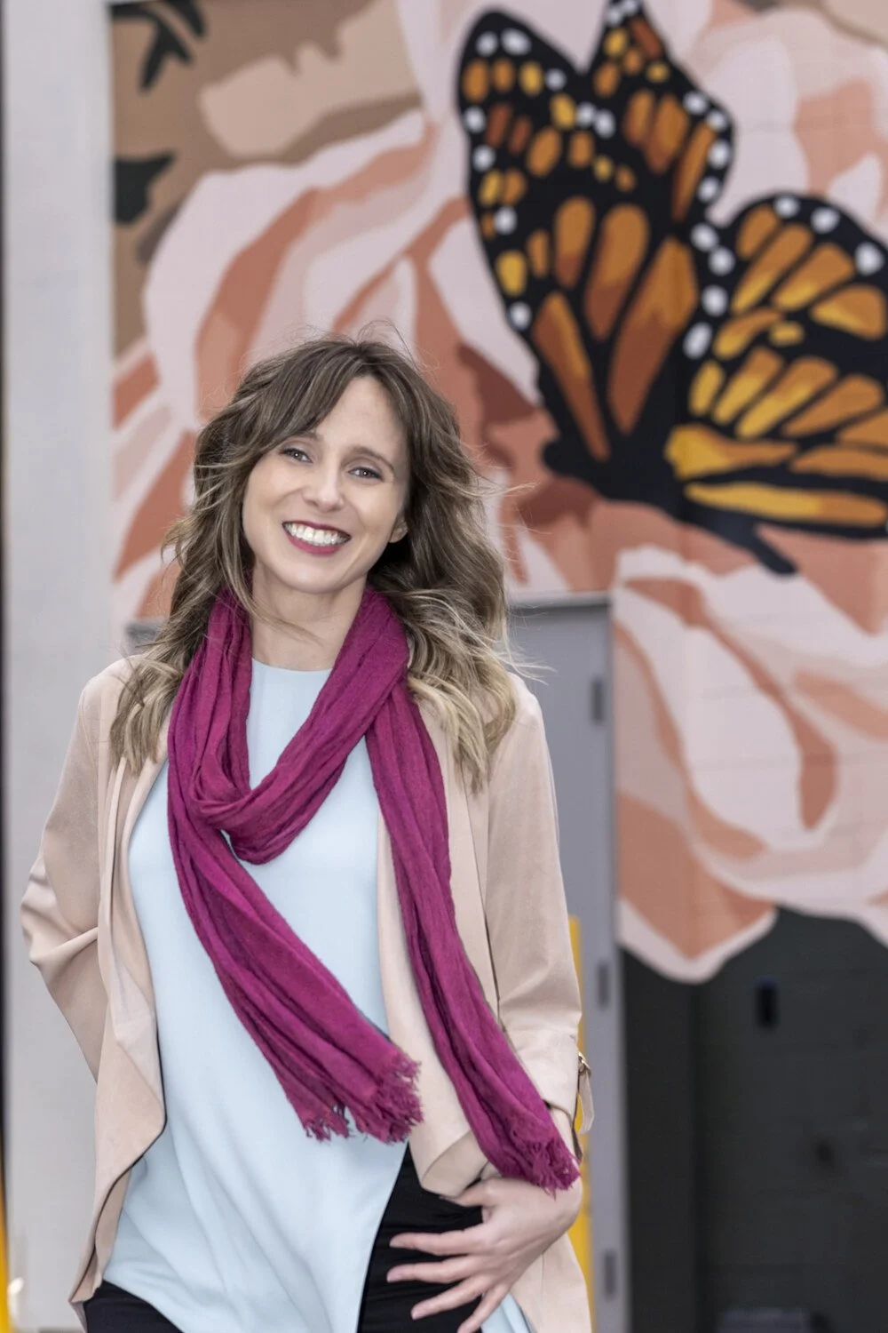 A woman with wavy, shoulder-length hair smiling at the camera. She is wearing a white top, a light beige blazer, and a magenta scarf. Behind her, there is a mural of a monarch butterfly on the wall.