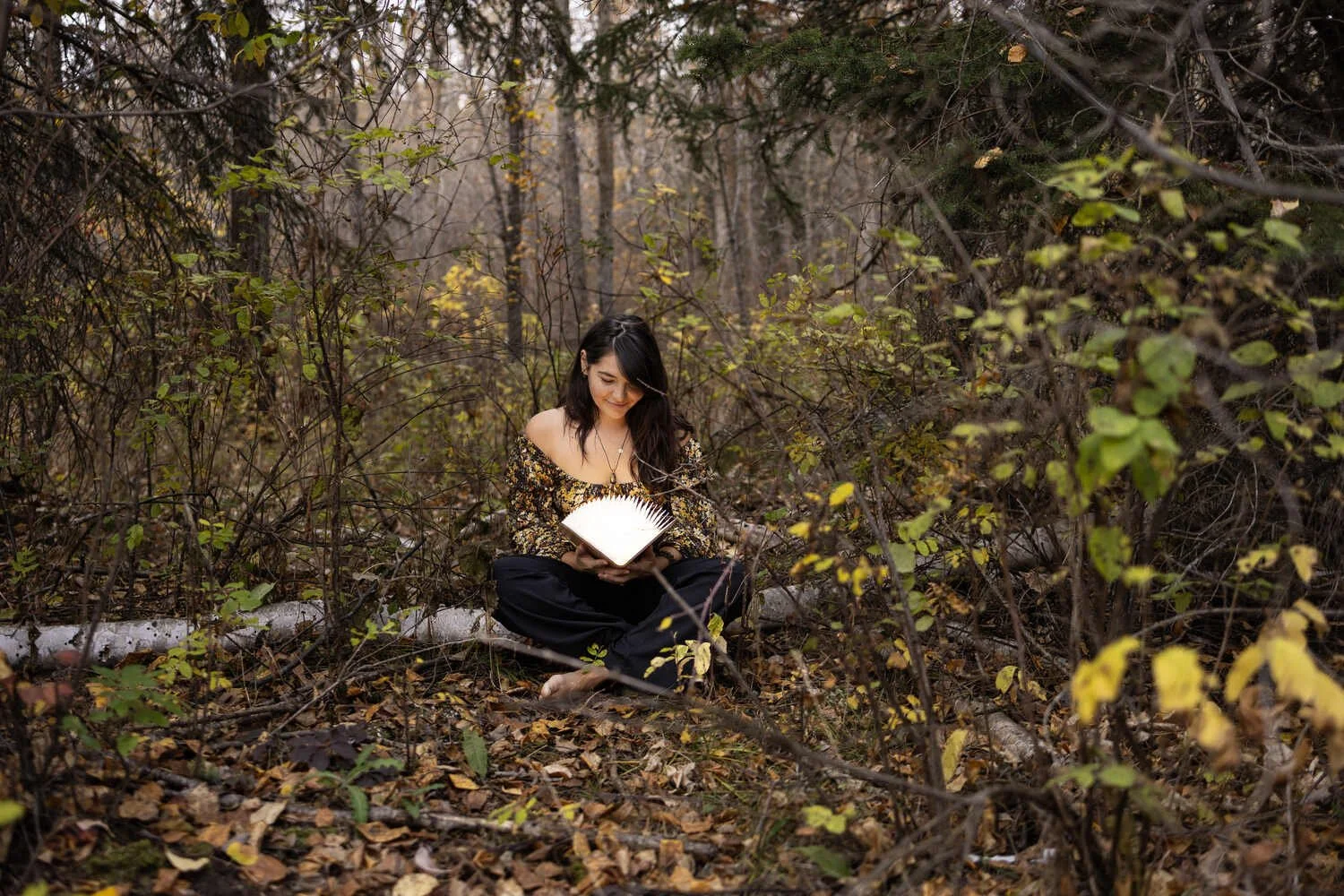 A woman with black hair sitting cross-legged on the forest floor, reading a white book, surrounded by leafless trees and fallen autumn leaves.