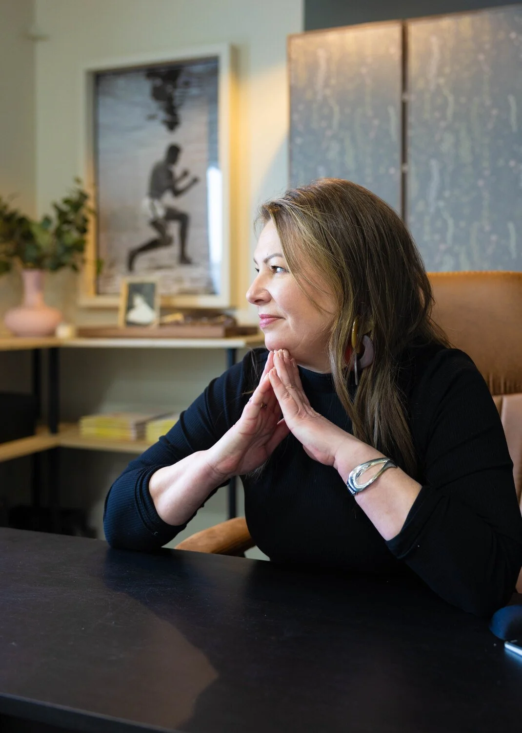 A woman with long brown hair, wearing a black top, sitting at a dark table with her hands clasped under her chin, looking to the side with a relaxed and contemplative expression in a modern room with artwork and plants in the background.