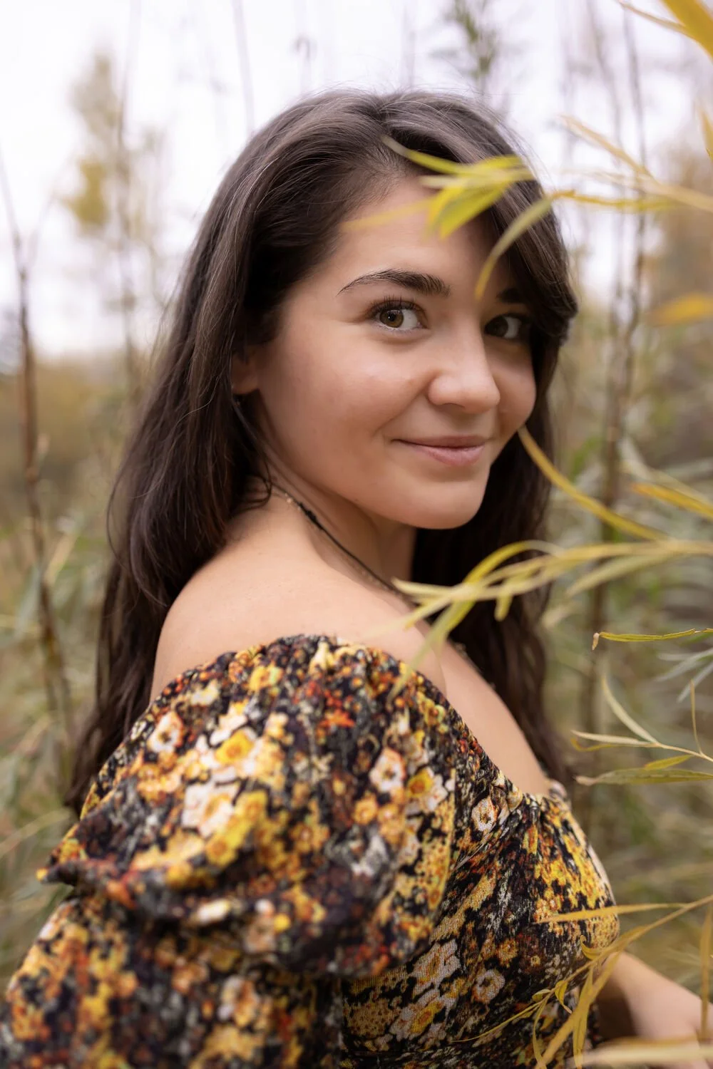 A woman with long dark hair smiling outdoors amidst yellowish-green plant leaves, wearing a floral dress.