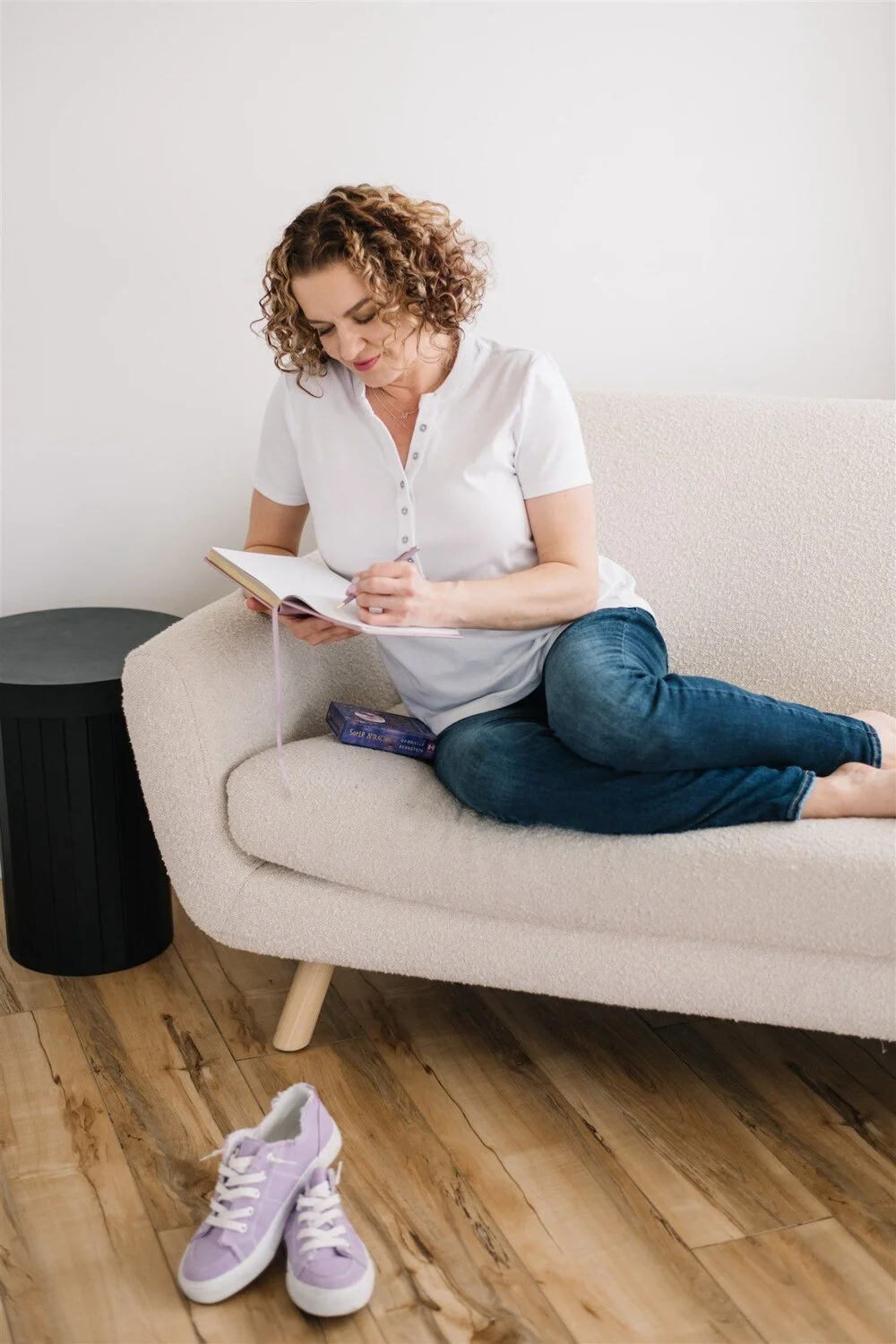 Photographer and coach Maggie Nowak sitting on a beige couch, reading a book, with purple sneakers on the floor in front of her and a black side table nearby.