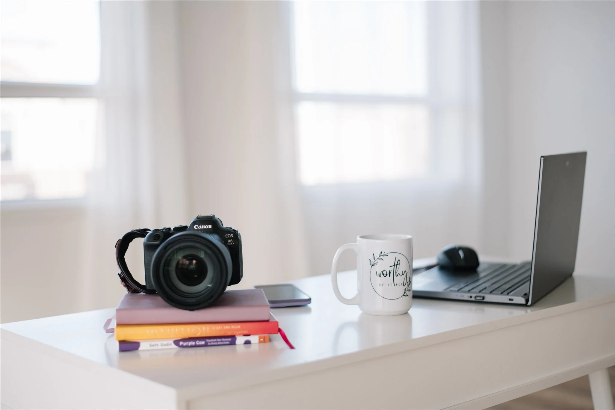 A white desk with a camera resting on a stack of books, a smartphone, a mug with the words "worthy" and floral design, a laptop, and a computer mouse in a bright room with white walls and large windows.