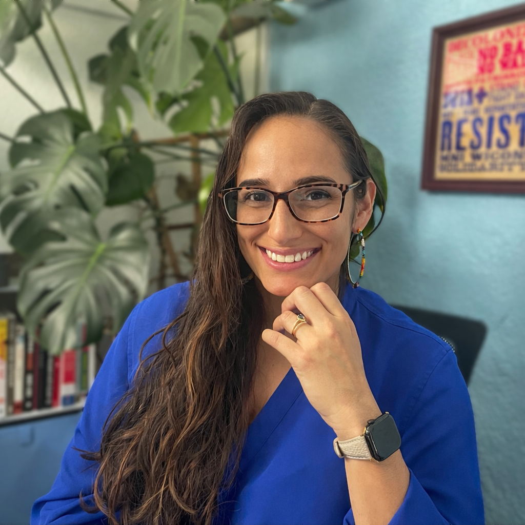 A woman with long wavy brown hair, wearing glasses, a blue top, and rainbow earrings, smiling while resting her chin on her hand in an indoor setting with a large leafy houseplant and a framed poster on the wall behind her.