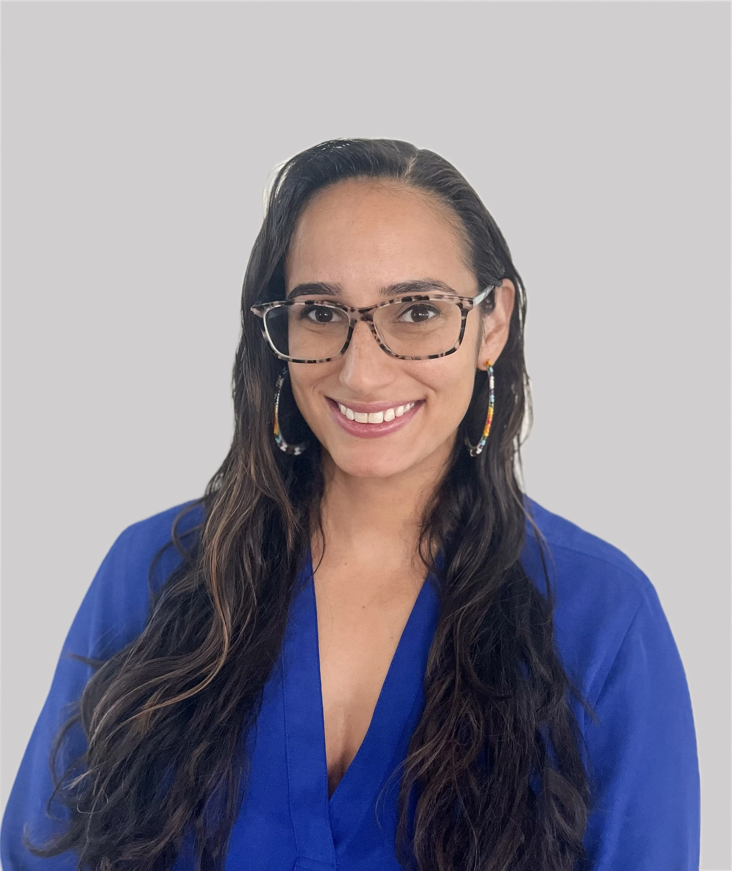 A woman with long dark wavy hair, wearing glasses, colorful earrings, and a blue top, smiling against a light gray background.