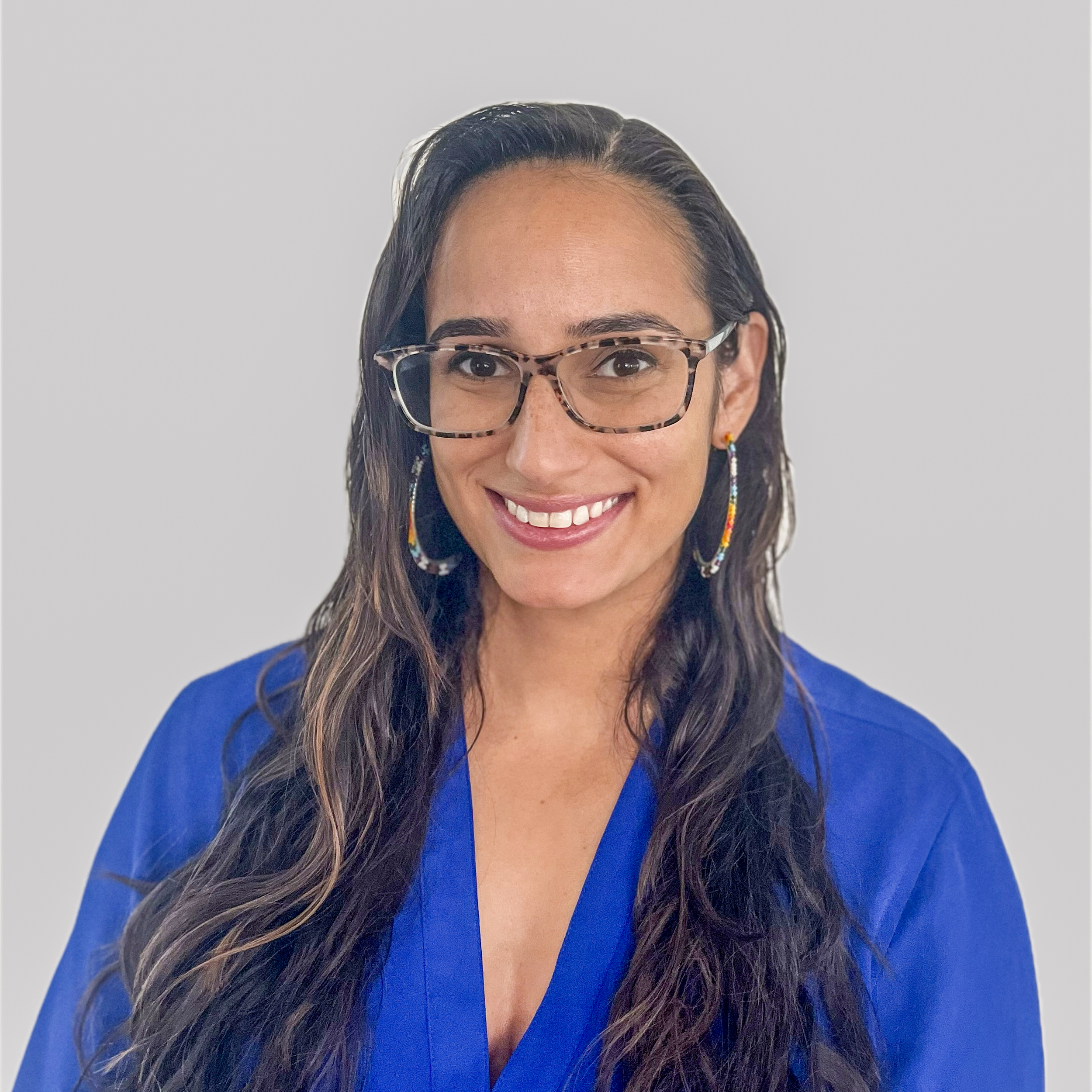 Smiling woman with long dark hair, wearing glasses, colorful earrings, and a blue top, against a plain light gray background.