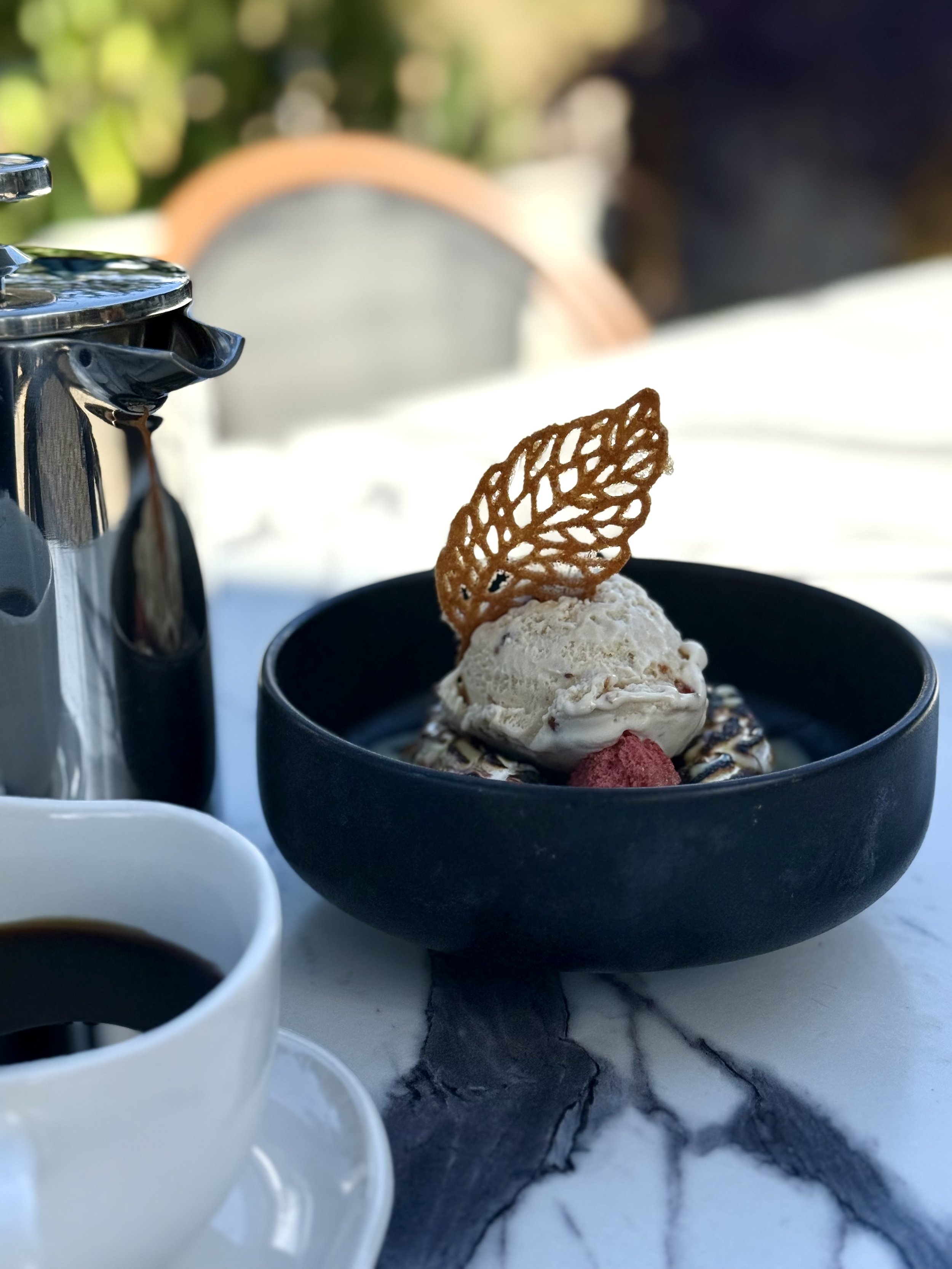 A bowl of ice cream with a decorative cookie leaf on top, served with a cup of coffee on a marble table outdoors.