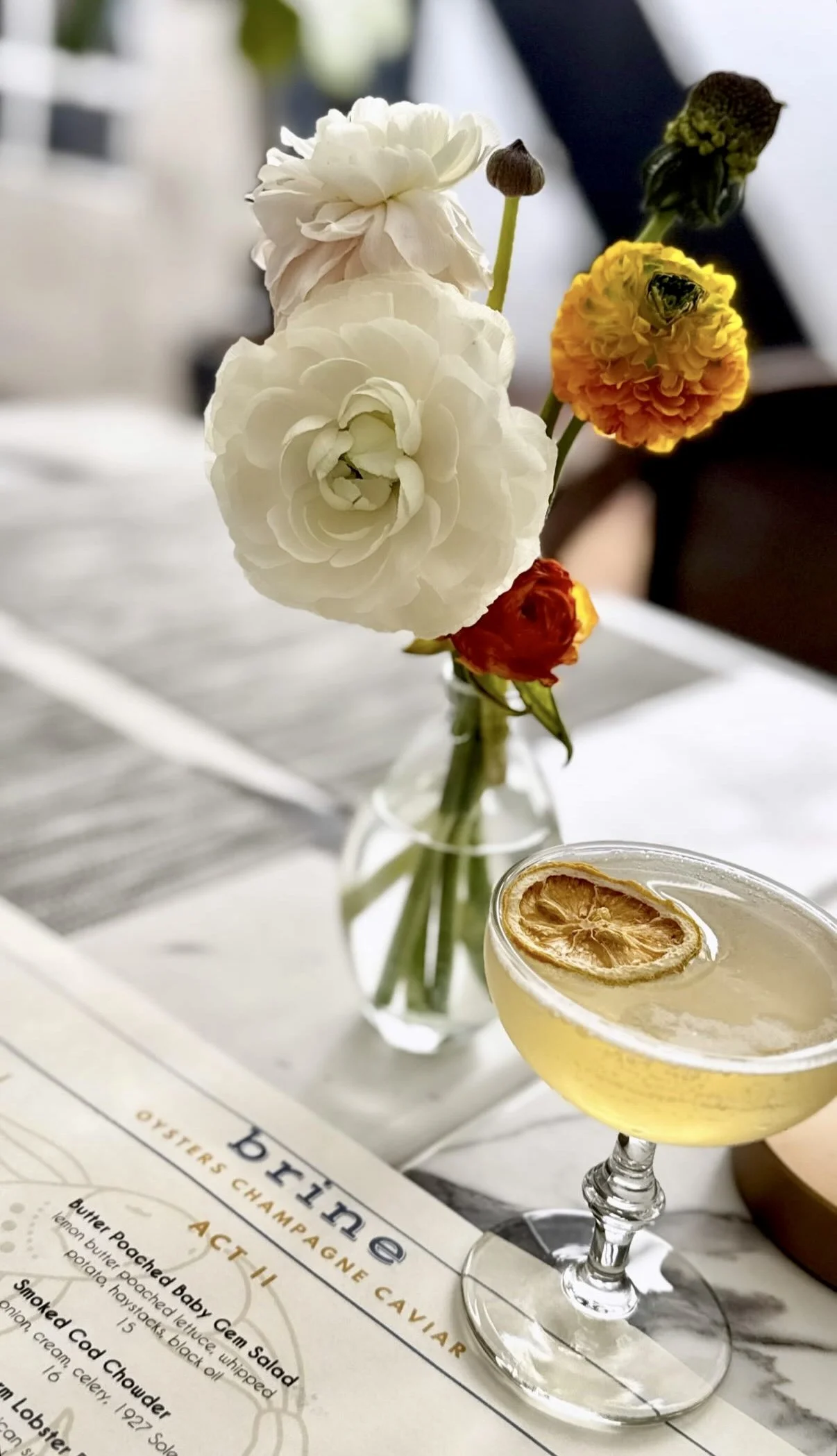 A close up of a table showing a small flower arrangement with white and orange flowers, and a cocktail with a dehydrated lemon wheel in a coupe glass.