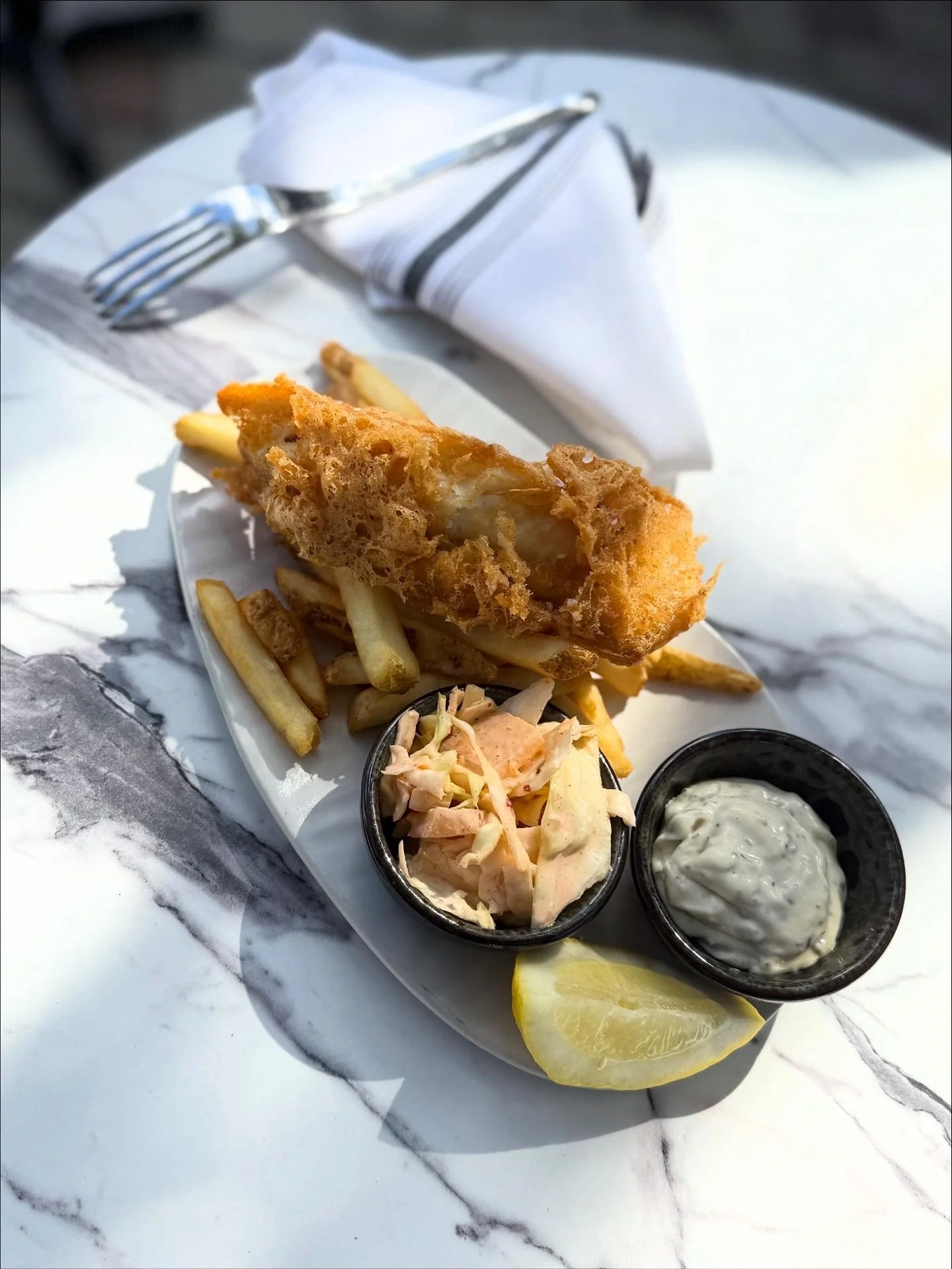 Fried cod fish fillet with French fries, coleslaw, tartar sauce, and a lemon wedge on a white platter, on a marble table.