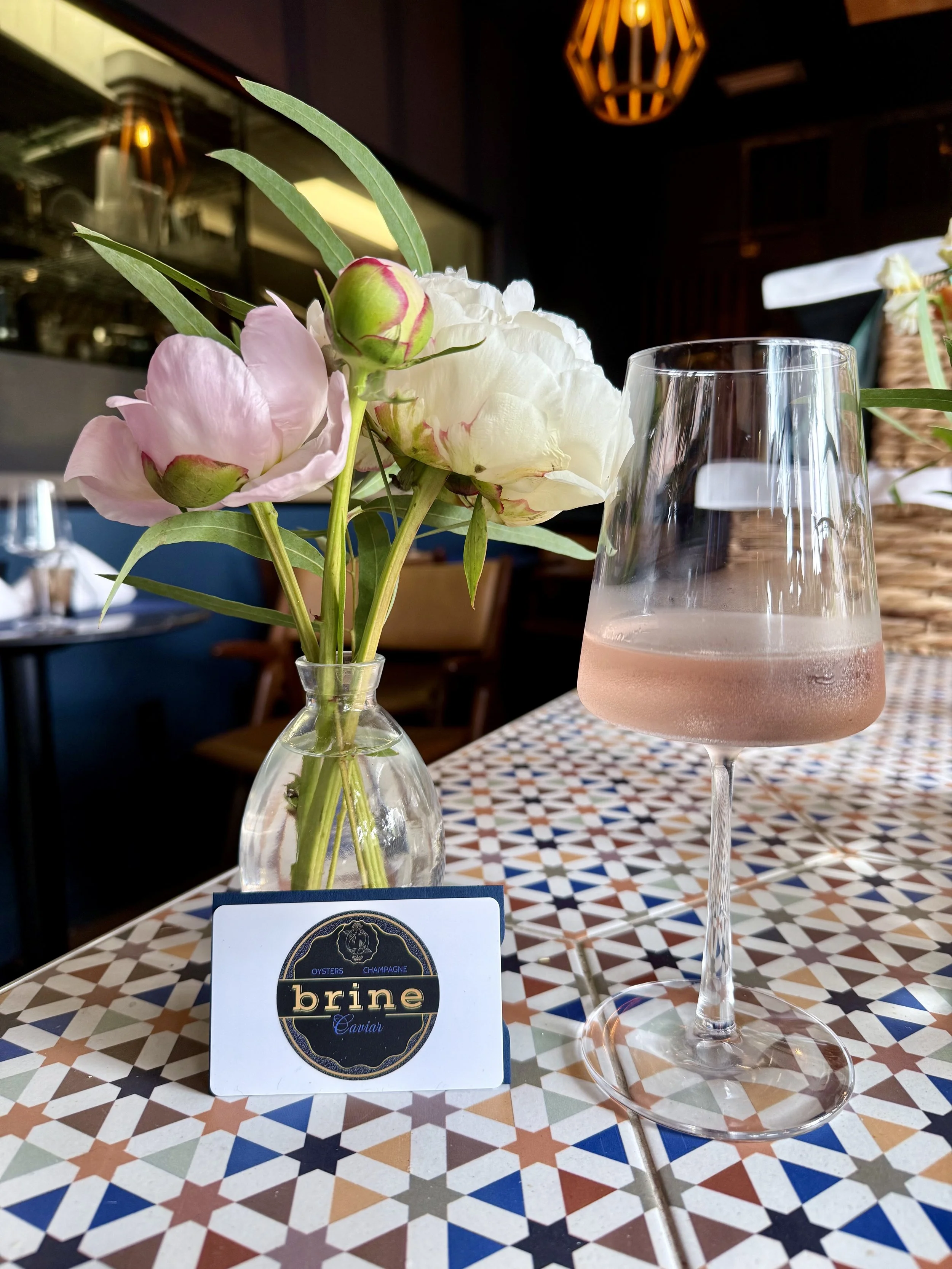 A vase with pink and white peonies and green leaves, a gift card reading 'brine caviar,' and a glass of sparkling rose wine on a patterned table in a restaurant setting.