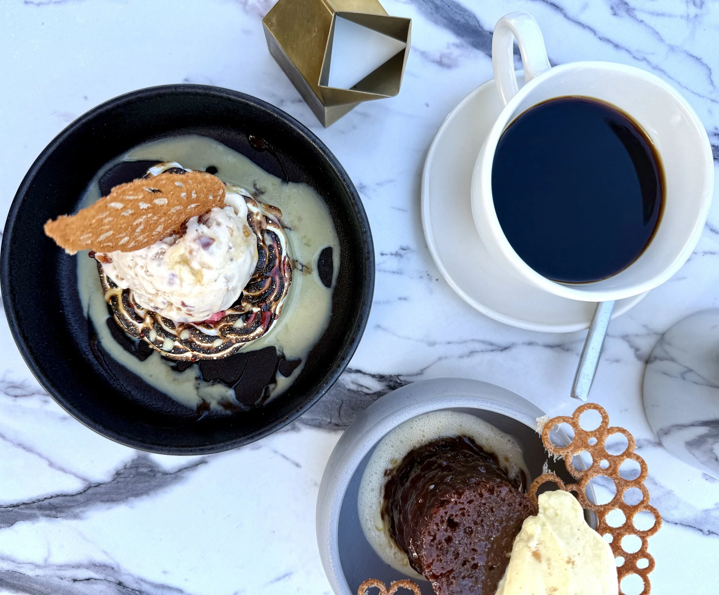 A white marble table with a cup of black coffee, a bowl of chocolate dessert with ice cream and cookie garnish, and a small dish with a chocolate-covered treat and pieces of cookies.