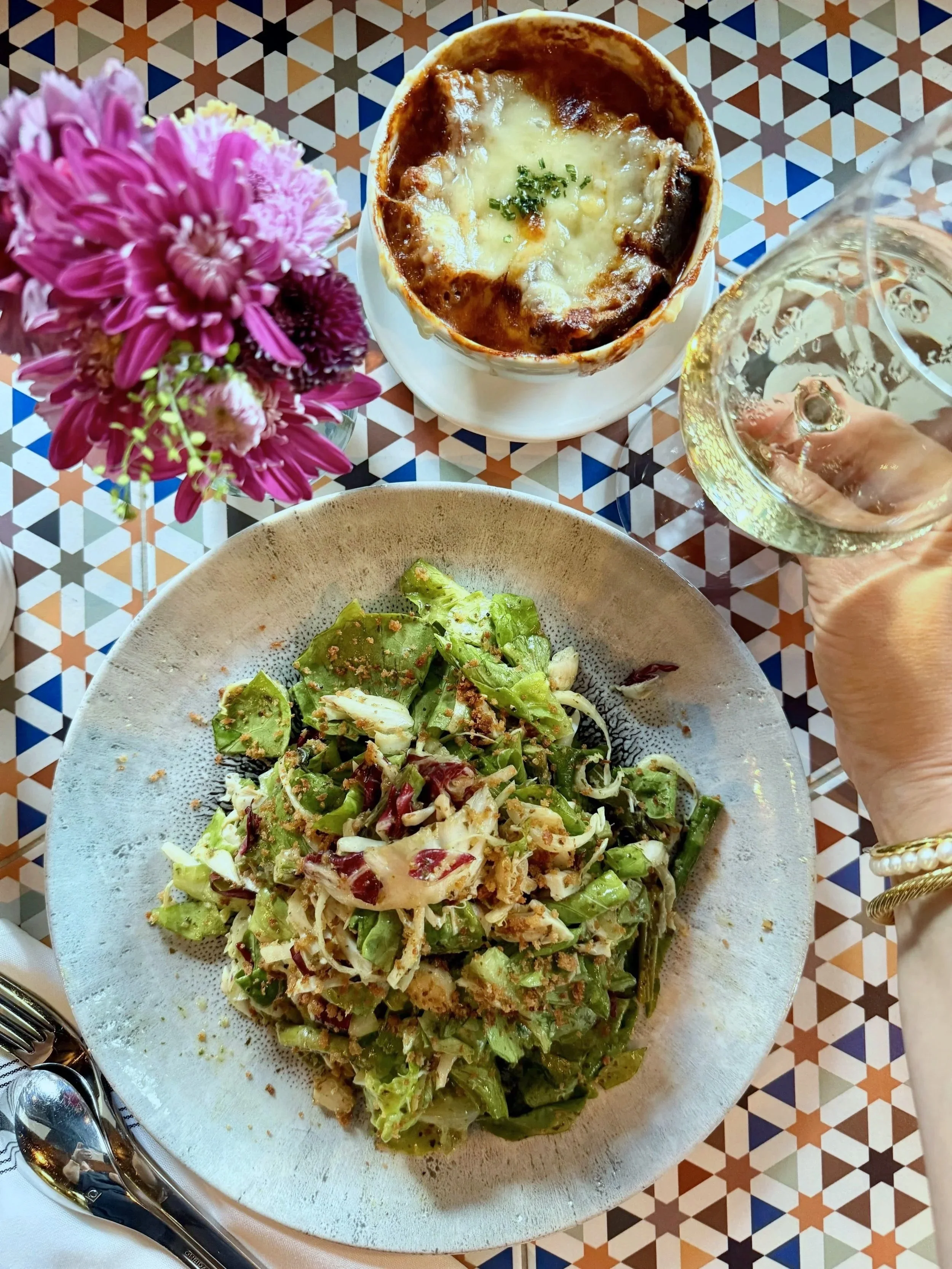 A plate of salad with chopped lettuce, radicchio, and lump crab meat and asparagus, alongside a small bowl of French onion soup topped with melted cheese, garnished with chopped herbs, and a glass of white wine on a colorful geometric tiled table.