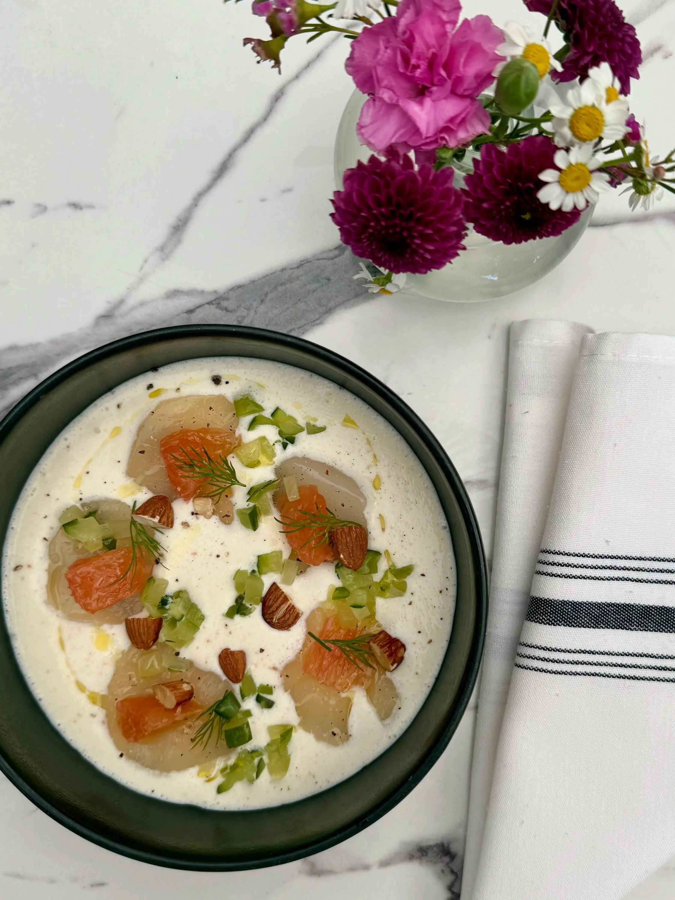 A bowl of cured fish, in a light cream sauce topped with trout roe, herbs, and nuts on a white marble table with a white napkin and a vase of pink, purple, and white flowers.