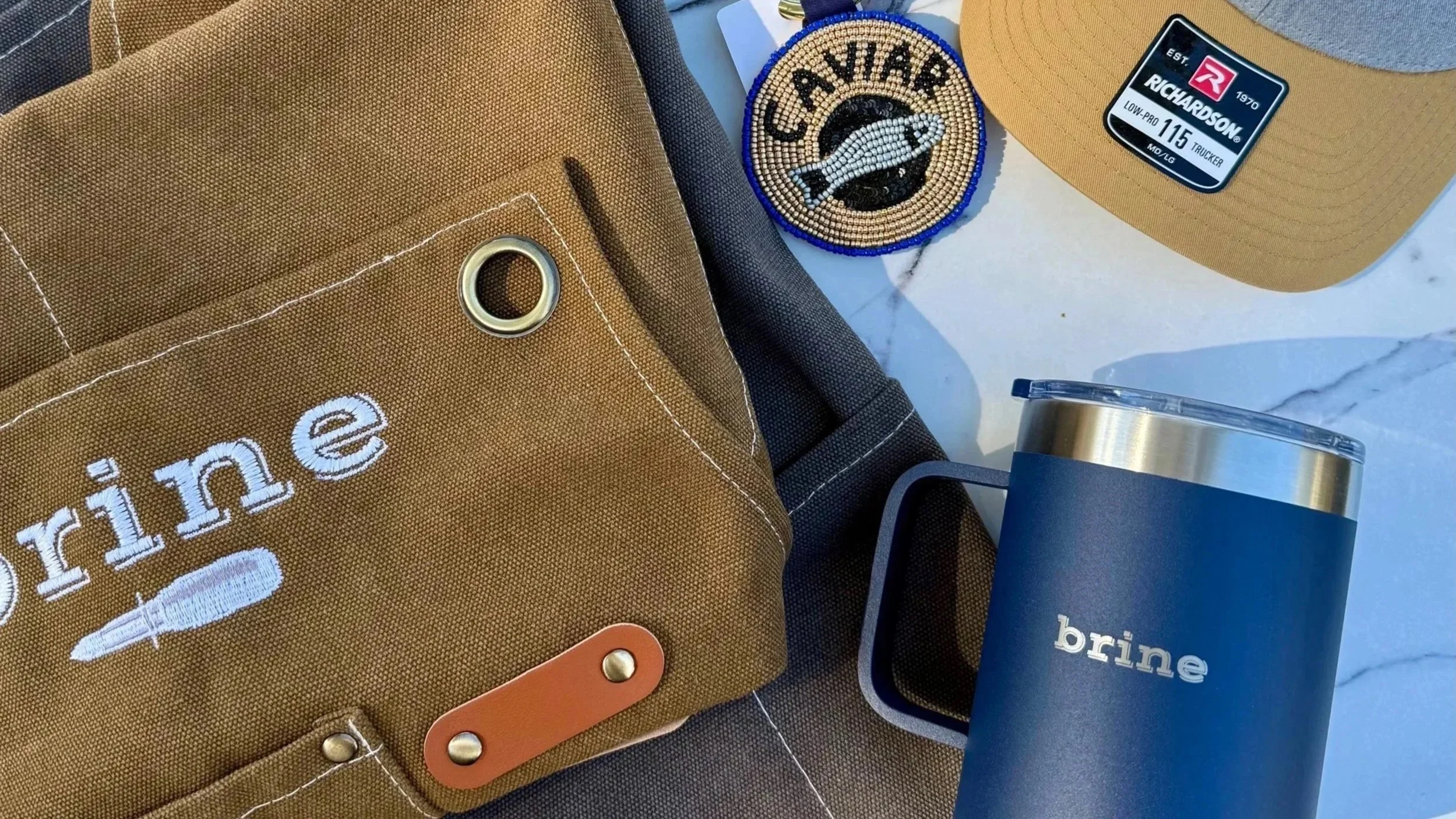 Banded merch including a tan canvas apron with embroidered 'brine' text, a beaded keychain with a fish and the word 'caviar,' a tan trucker hat with embroidered 'brine' text, a navy blue tumbler labeled 'brine,' and a gray cap on a marble surface.