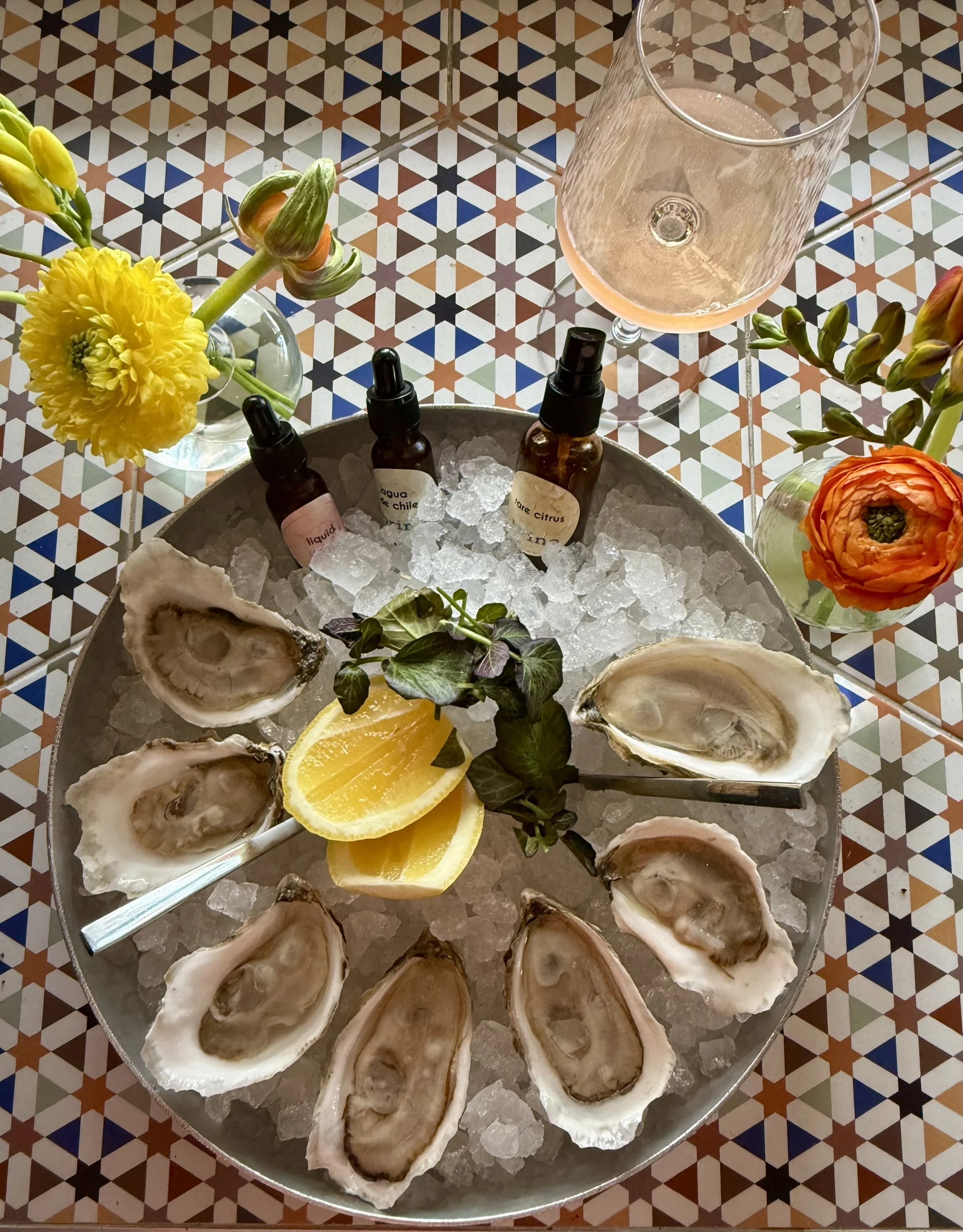 Oyster platter with ice, lemon wedges, and herbs, surrounded by flowers and spray bottles, with a glass of rosé wine on a patterned table.