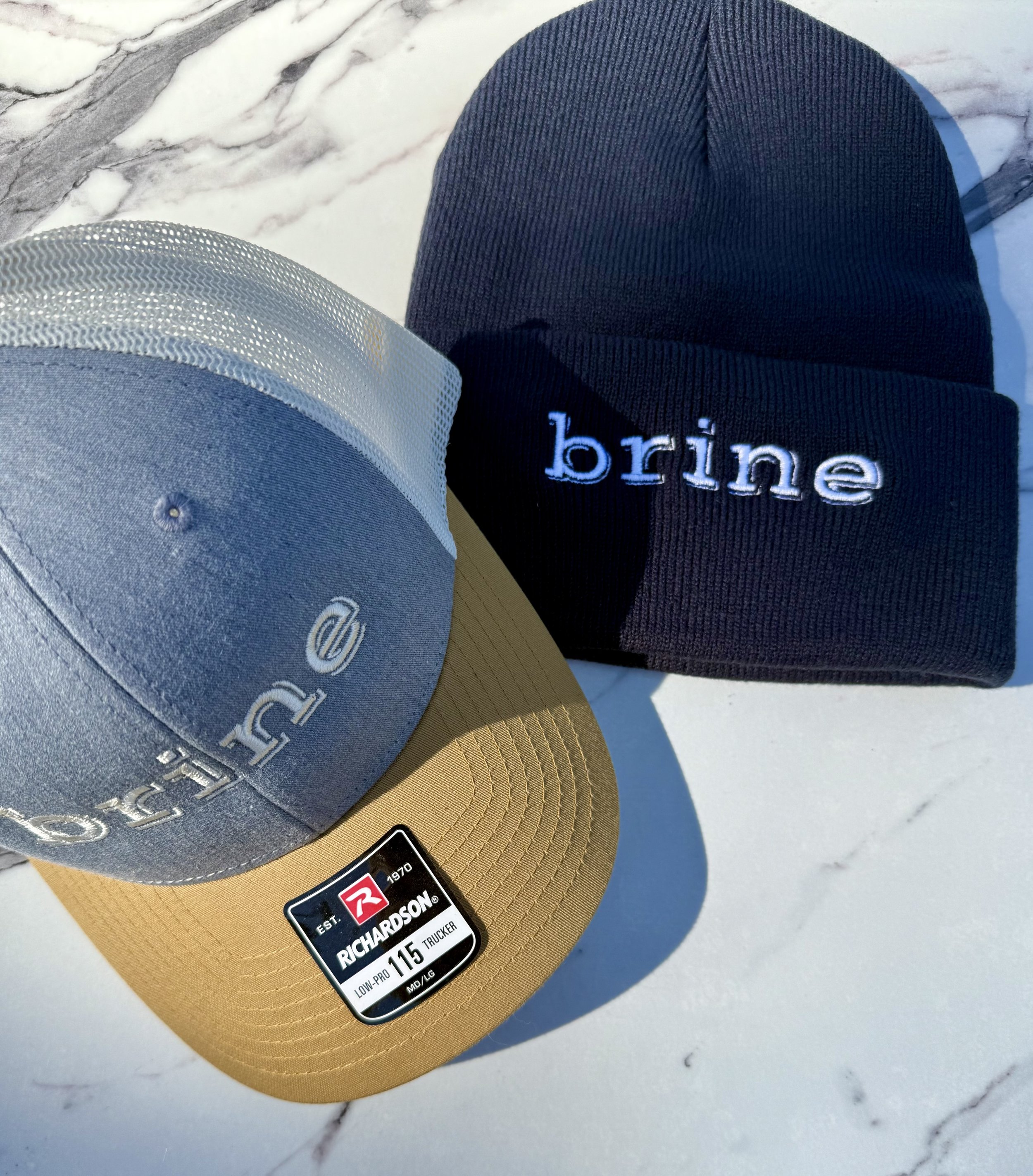 A gray and tan Richardson trucker cap with the word 'brine' emboidered in white and a navy knit beanie with the word 'brine' embroidered in white on a white marble surface.