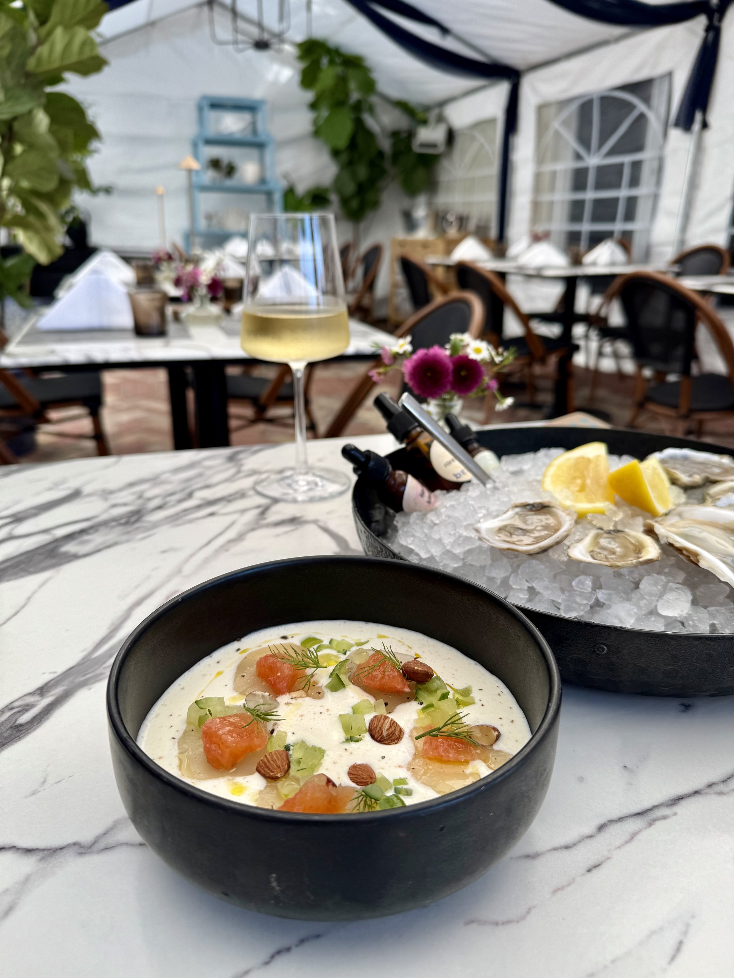 A seafood dish with cured fish in a light colored sauce, topped with orange trout roe, next to a dish of oysters and lemon wedges on ice, and a glass of white wine on a marble table at a restaurant with decorated tables and chairs.