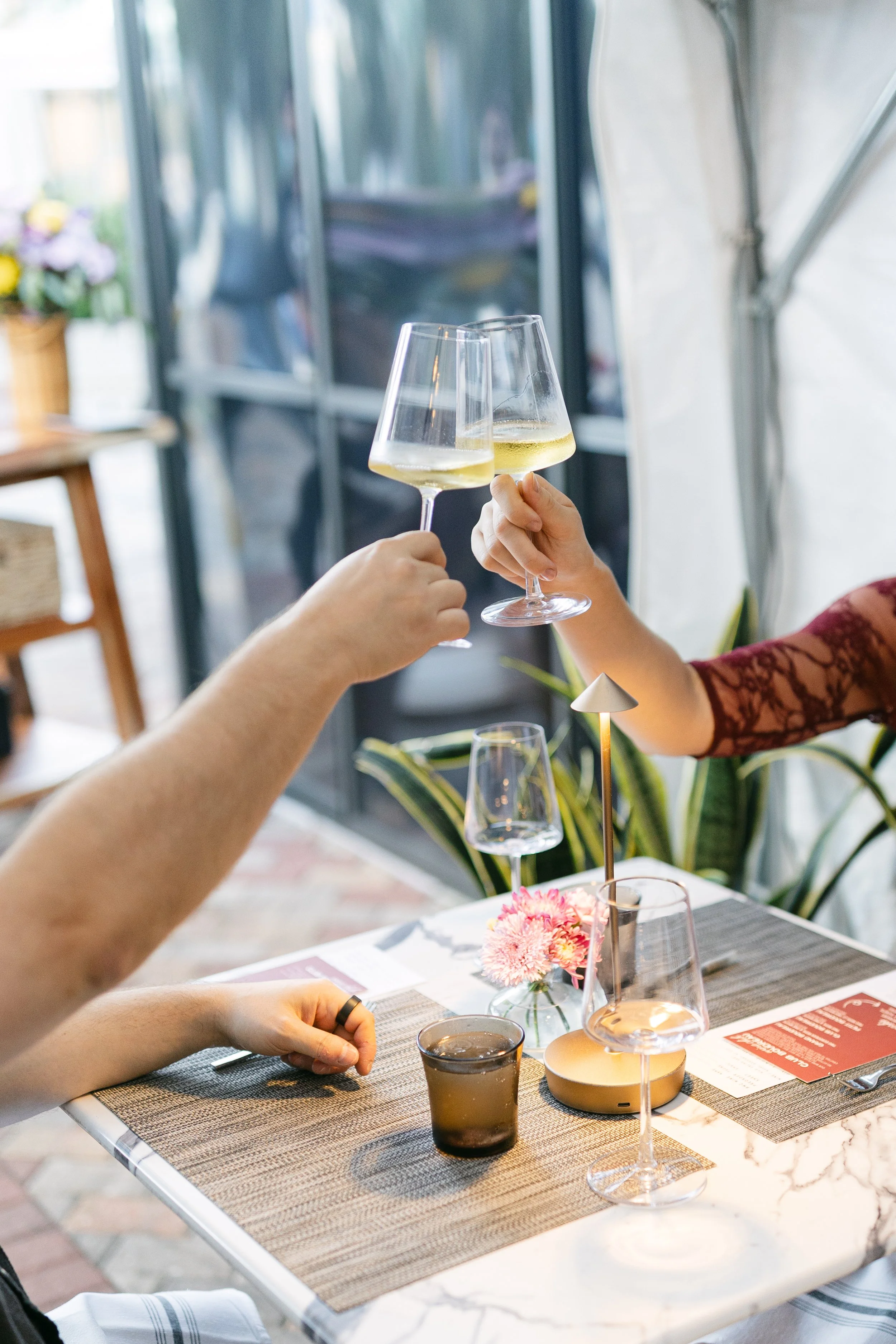 Two people clinking glasses of white wine at a restaurant table decorated with flowers, with a person sitting and a hand resting on the table.