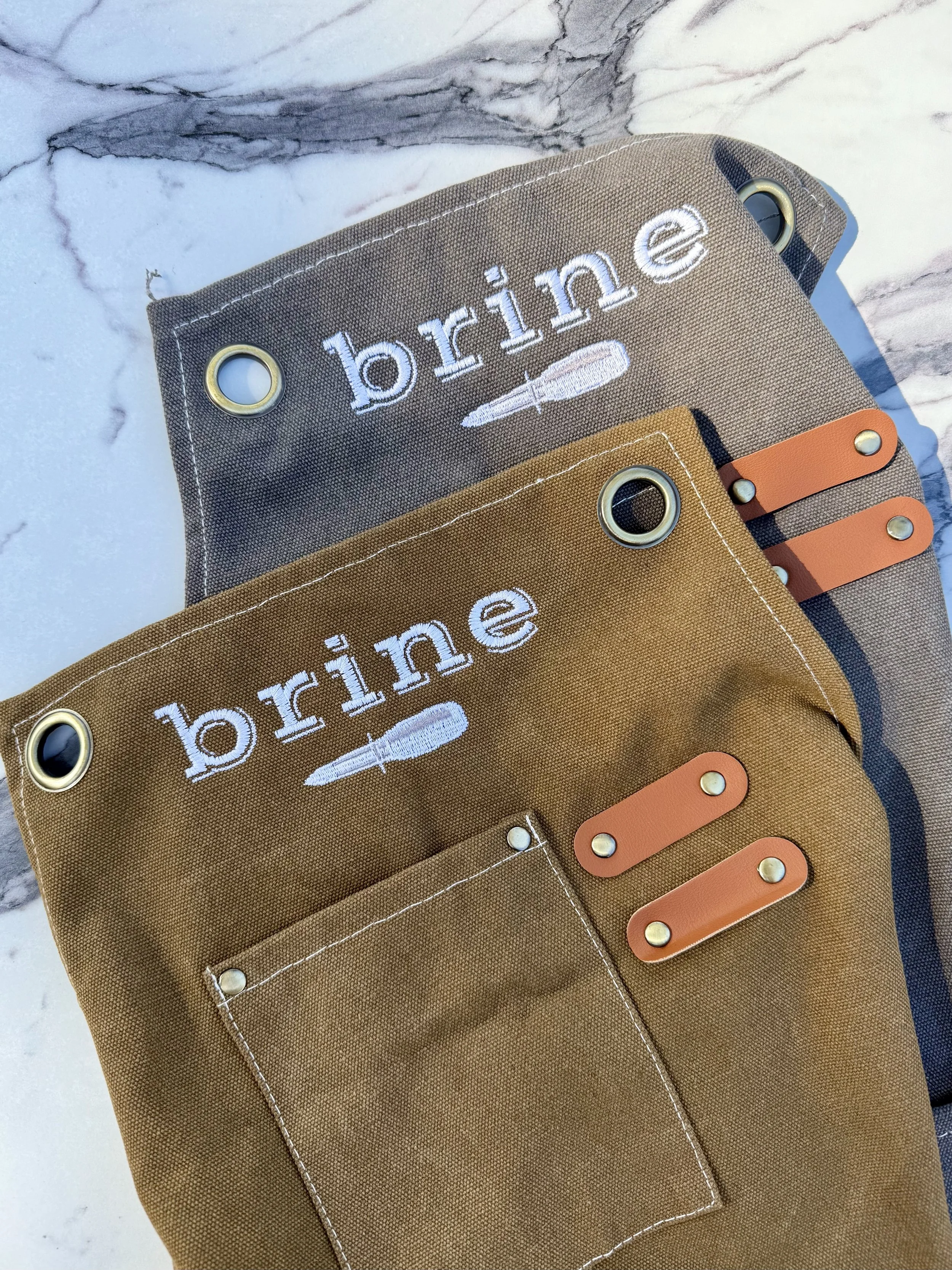 Two canvas aprons with the word 'brine' embroidered on them, featuring an oyster knife graphic, placed on a marble surface. One apron is grey, and the other is brown, each with metal grommet holes and brown leather straps.