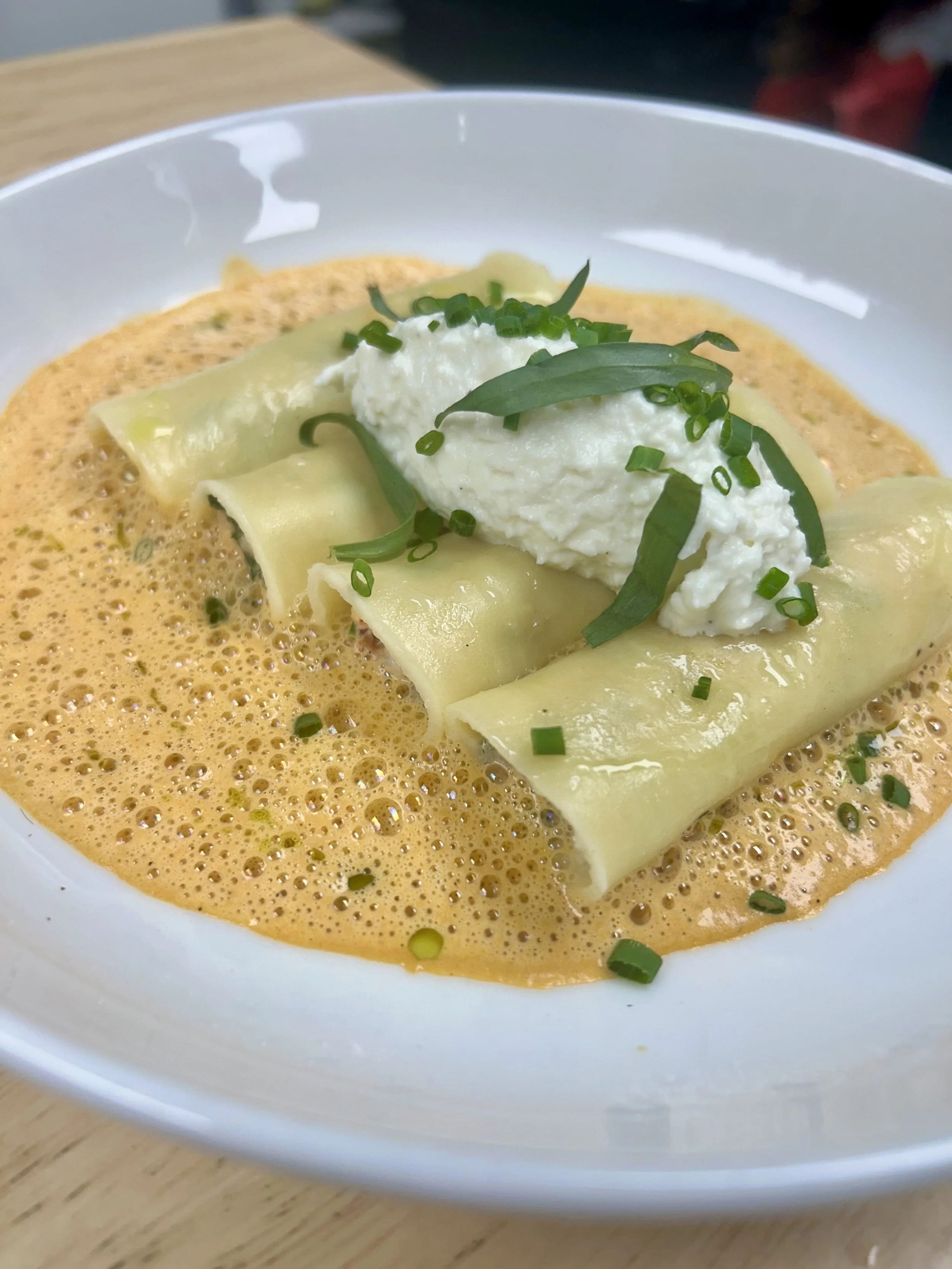 Plate of cheese tortellini filled with lobster, in a creamy orange sauce, topped with a dollop of ricotta cheese, chopped chives, and basil leaves.