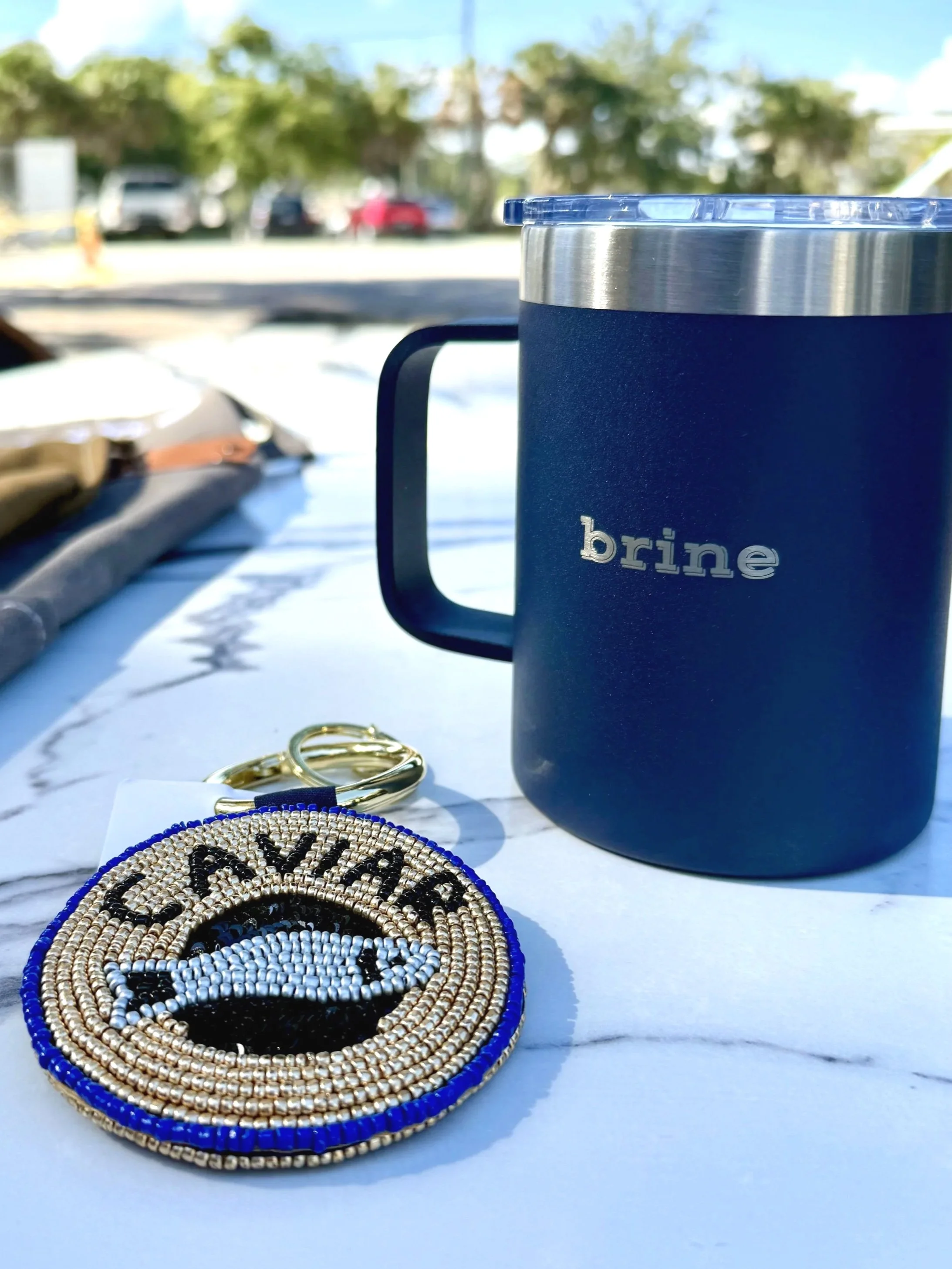 A blue brine travel mug, a beaded keychain with a fish and the word 'CAVIAR' on a white marble surface outdoors with trees and parked cars in the background.