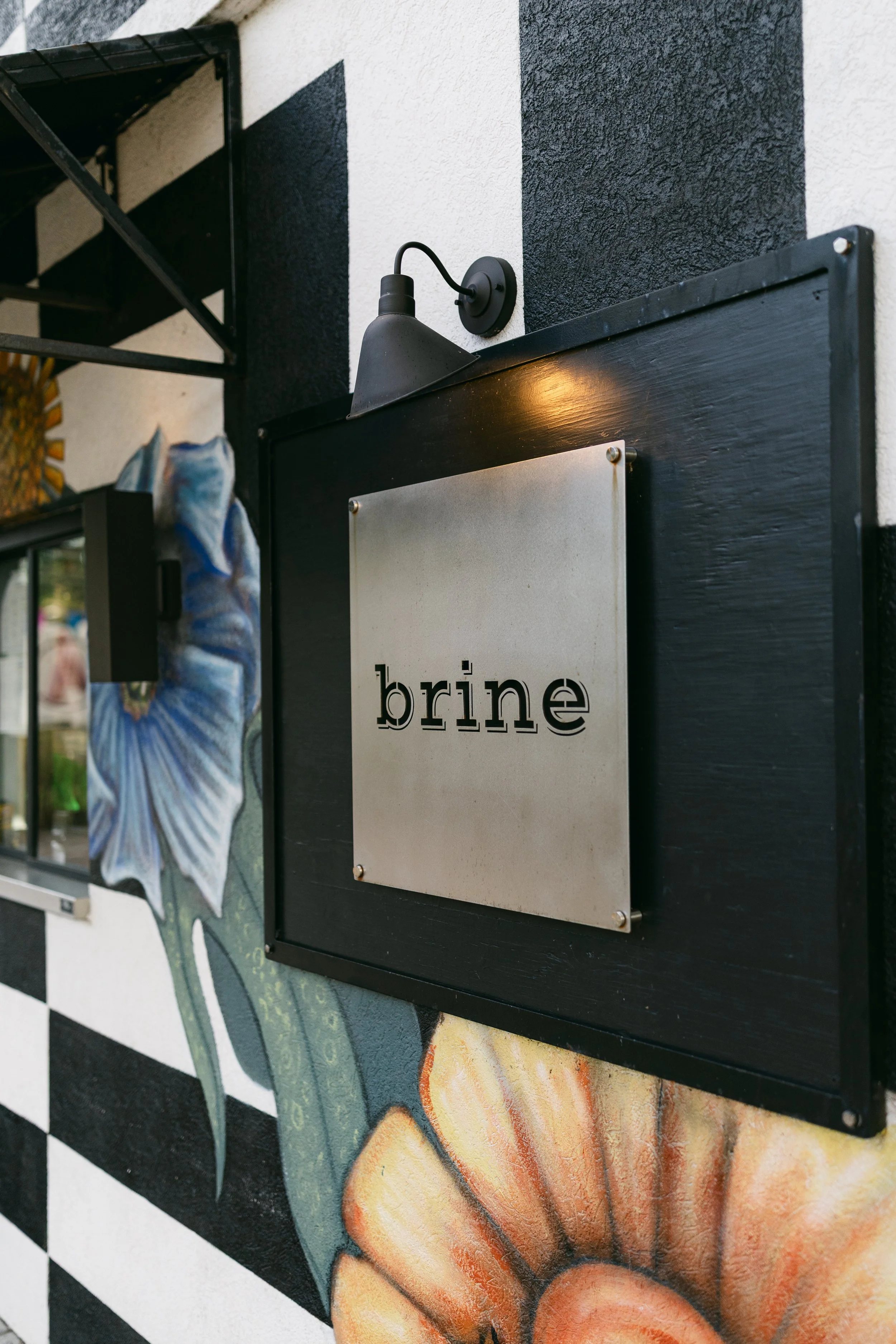 A photo of the restaurant's metal sign with their logo laser cut with the word 'brine'. The sign is on a black and white painted building with large blue and orange flowers