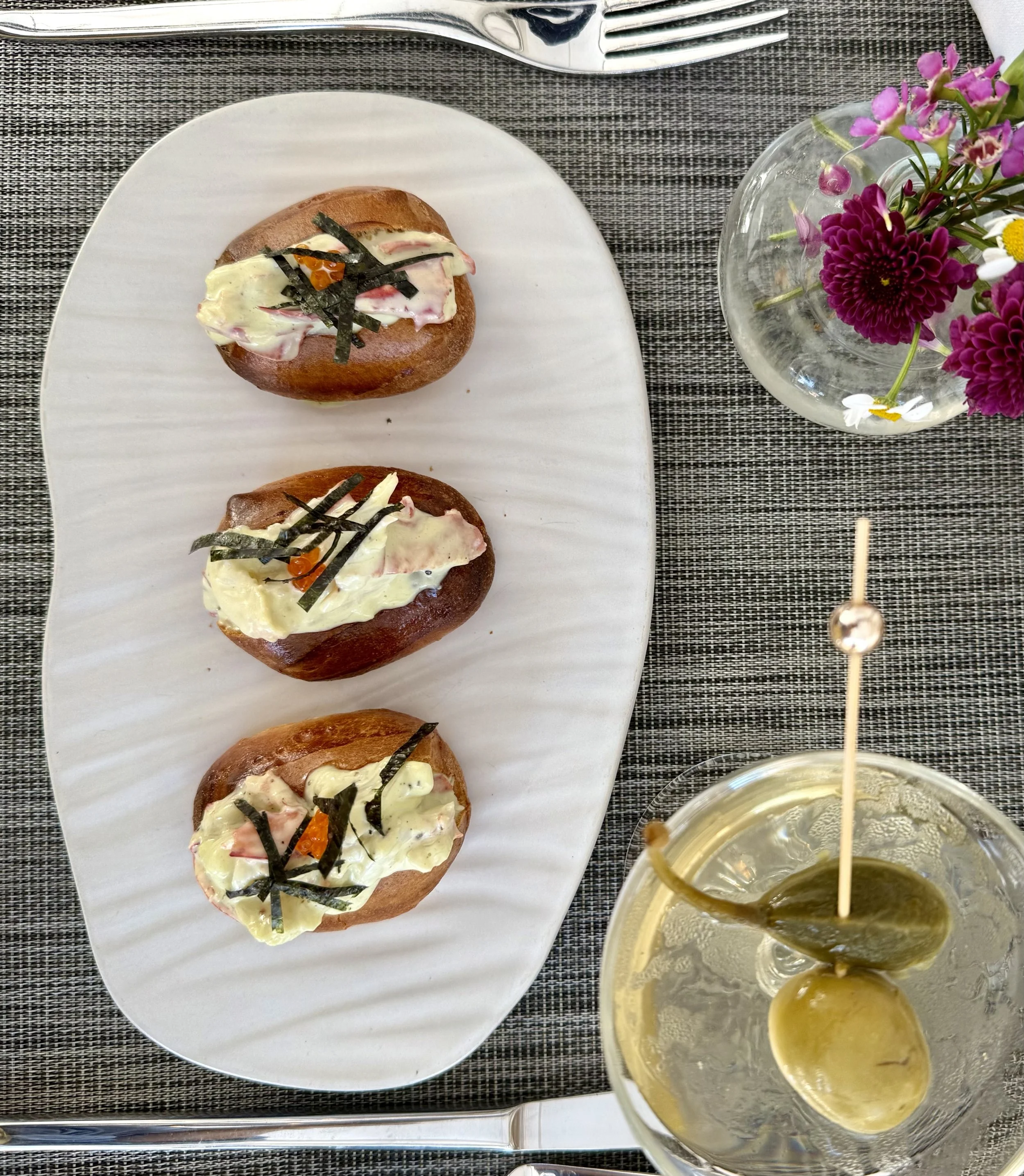 Three lobster roll sliders topped with mayonnaise, shredded nori, and trout roe on a white oval plate, with a fork above, a vase with pink and purple flowers to the right, and a drink with olives to the bottom right, all on a gray textured placemat.