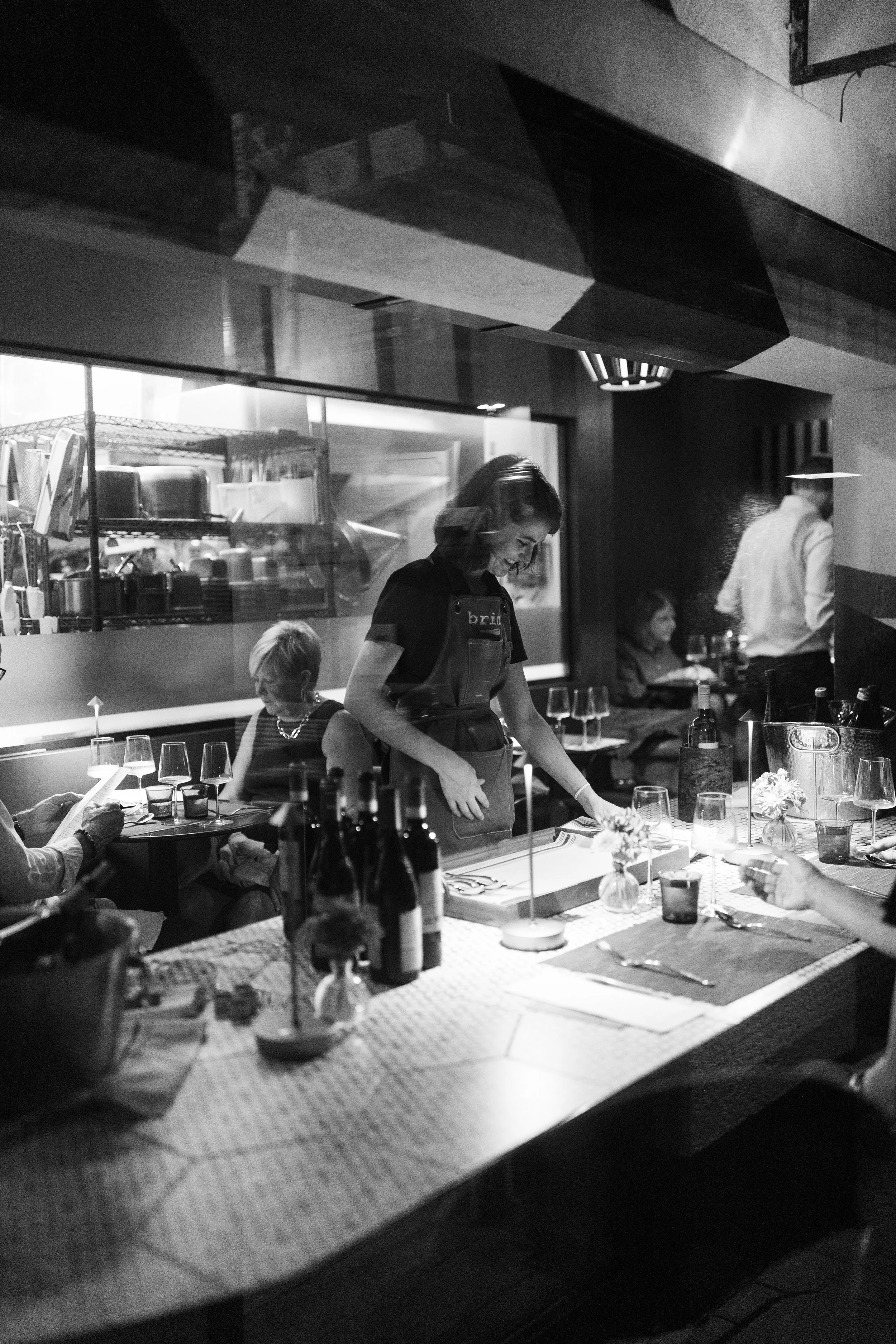 A black and white photo depicting a server working at a busy counter area with wine bottles, glasses and a placemat.