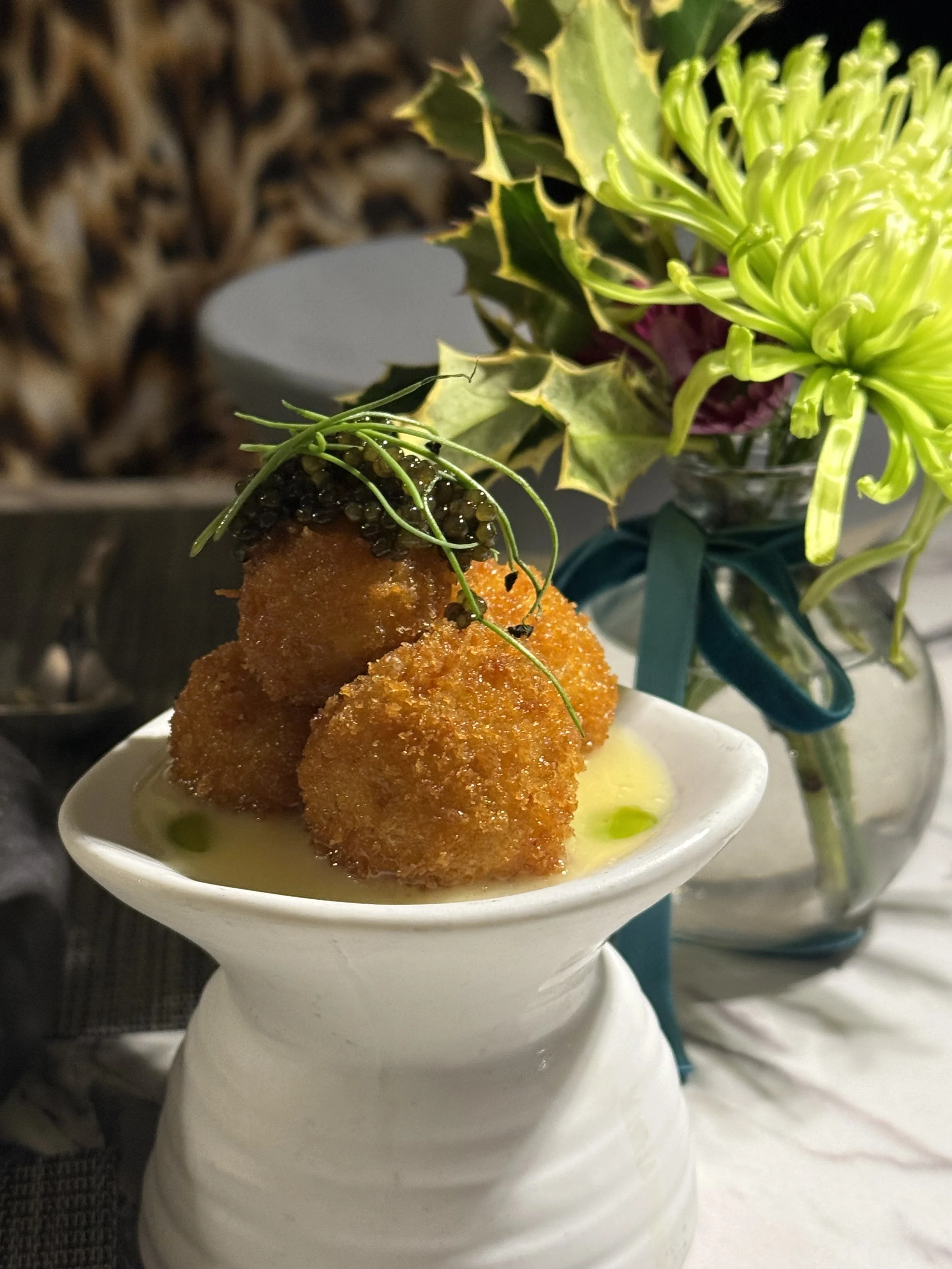 Fried appetizer balls garnished with herbs, served on a white plate with dipping sauce, and placed near a vase of flowers with green and purple blossoms.