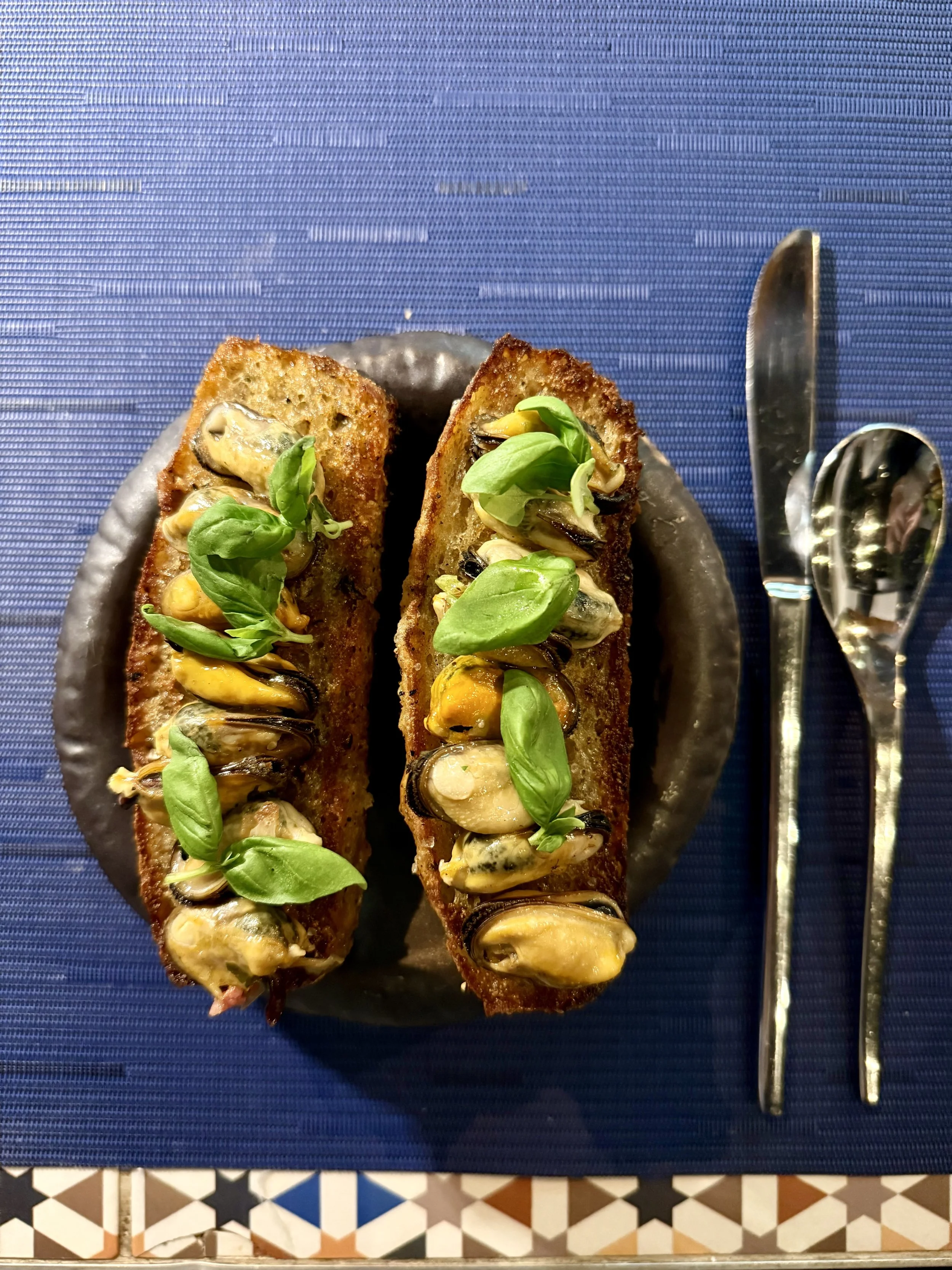 Two toasted baguette slices topped with mussels, basil leaves, and a drizzle of sauce on a dark plate, with a knife and spoon on a blue placemat.
