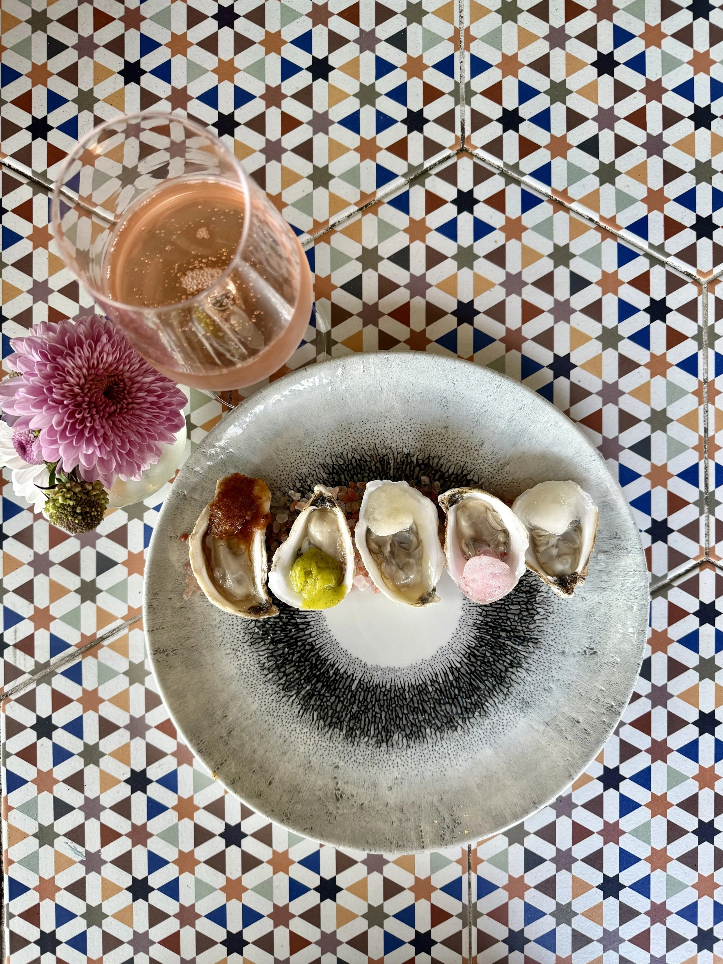An artful shot of 5 oysters lined up on a plate, each topped with a different color sorbet. A small purple flower arrangement and a glass of rose wine are also in the photo.