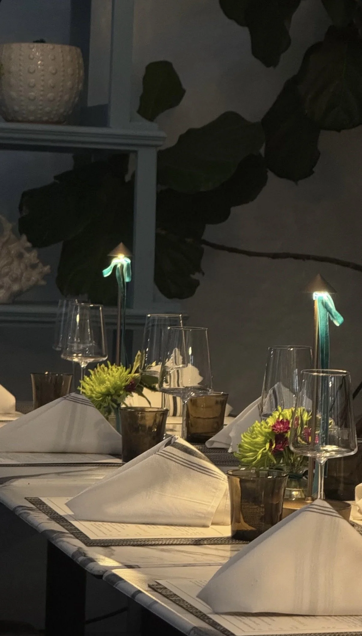 A low-lit photo that conveys a romantic and intimate atmosphere, with a table set with napkins, small arrangements of green and purple flowers, and table lamps. 