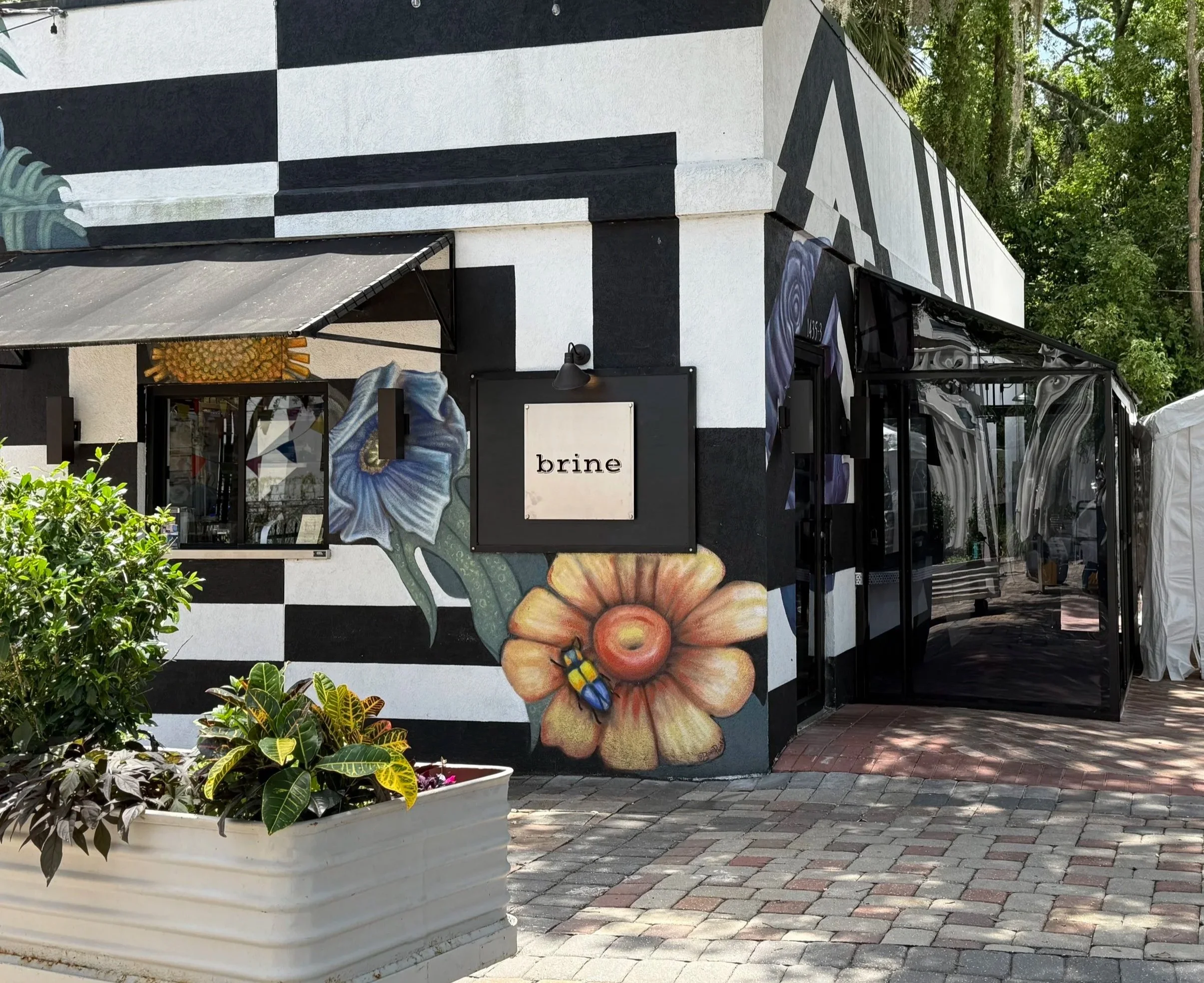 Exterior of a building with black and white painted stripes and floral murals, including a large flower, a bee, and a blue flower. The building has a sign with the word 'brine' and an outdoor seating area with glass doors and greenery.