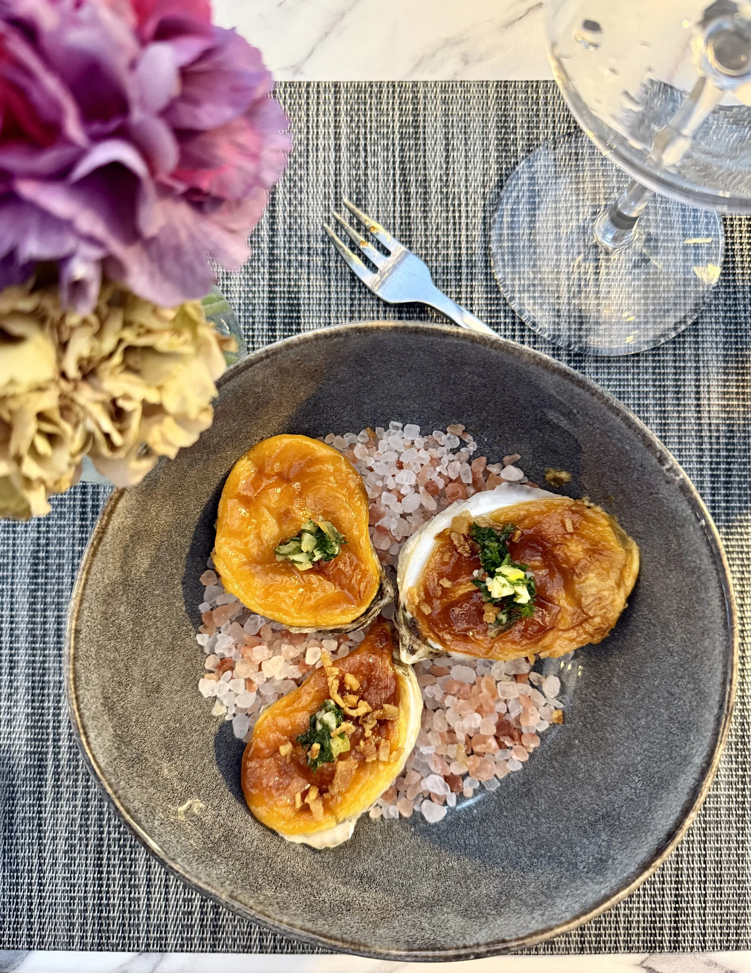 A black plate with three halved baked oysters topped with herbs and garnished with chopped nuts, set on a salt bed. The plate is on a textured gray placemat with a fork, a glass of water, and a floral arrangement nearby.