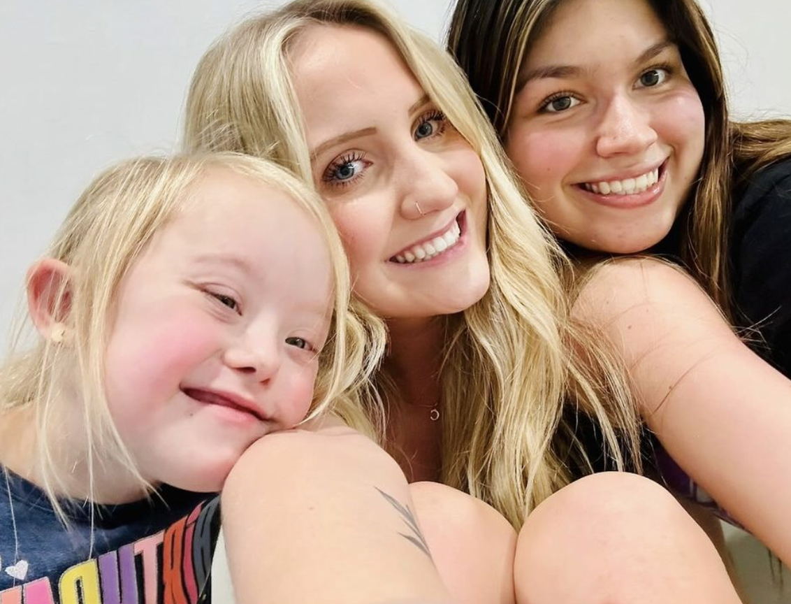 Three smiling women and a young girl taking a close-up selfie together indoors.