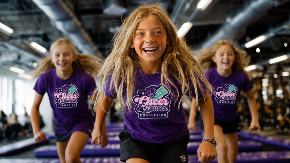 Three girls with long blonde hair and purple shirts running excitedly in a gym or dance studio.