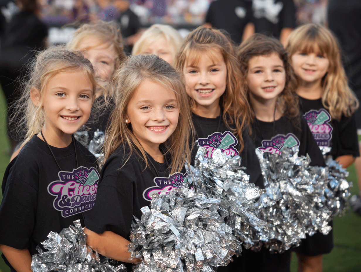 Group of young girls holding silver pom-poms, smiling, wearing black cheerleading shirts with a logo that says 'The Cheer & Dance Connection'.
