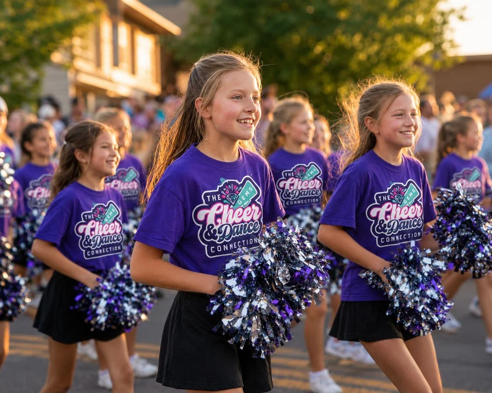Young cheerleaders in purple shirts and black skirts holding pom-poms and smiling during a parade or outdoor event.