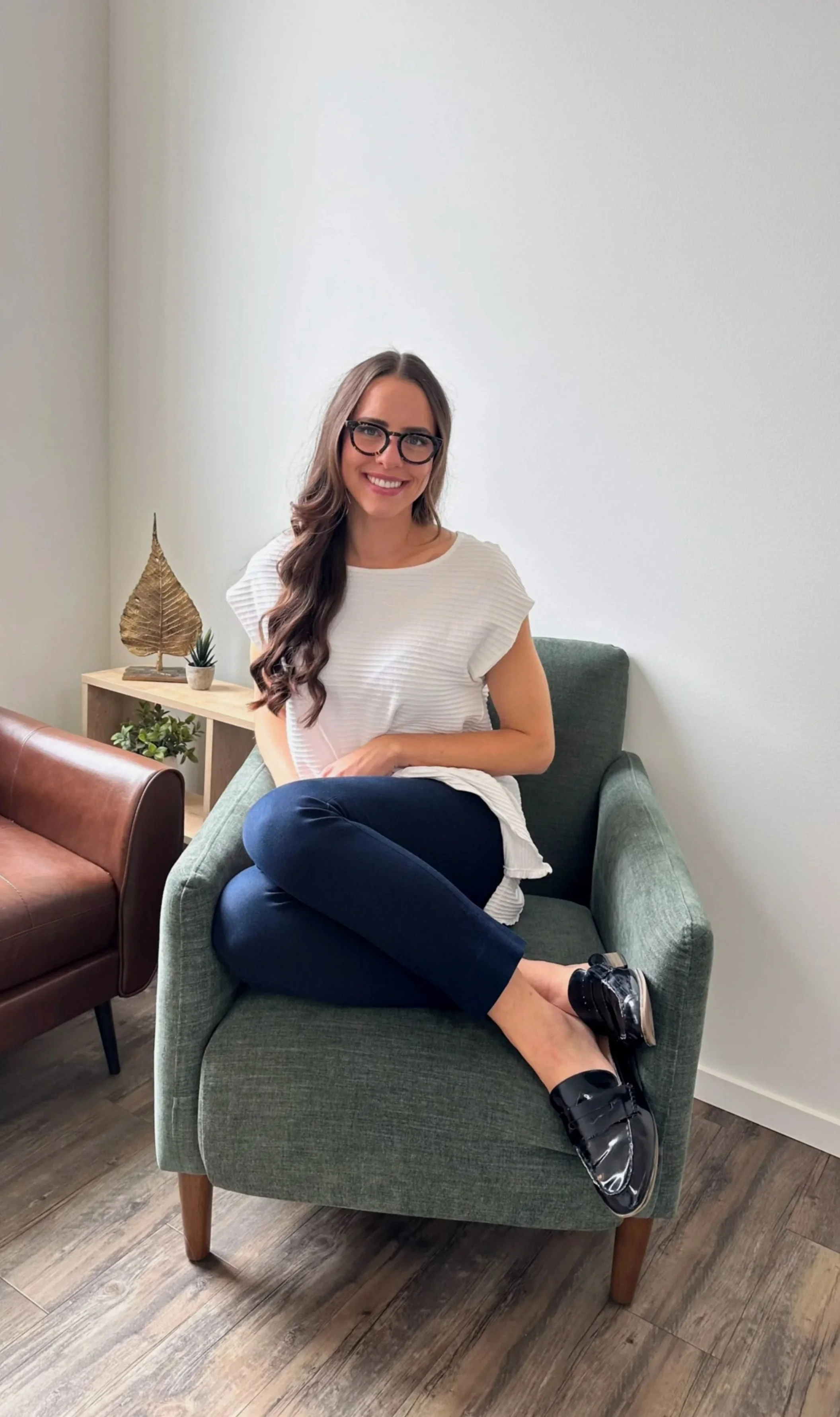 A woman with long brown hair, glasses, wearing a white top, dark pants, and black shoes, sitting on a green armchair with her legs crossed, smiling at the camera.