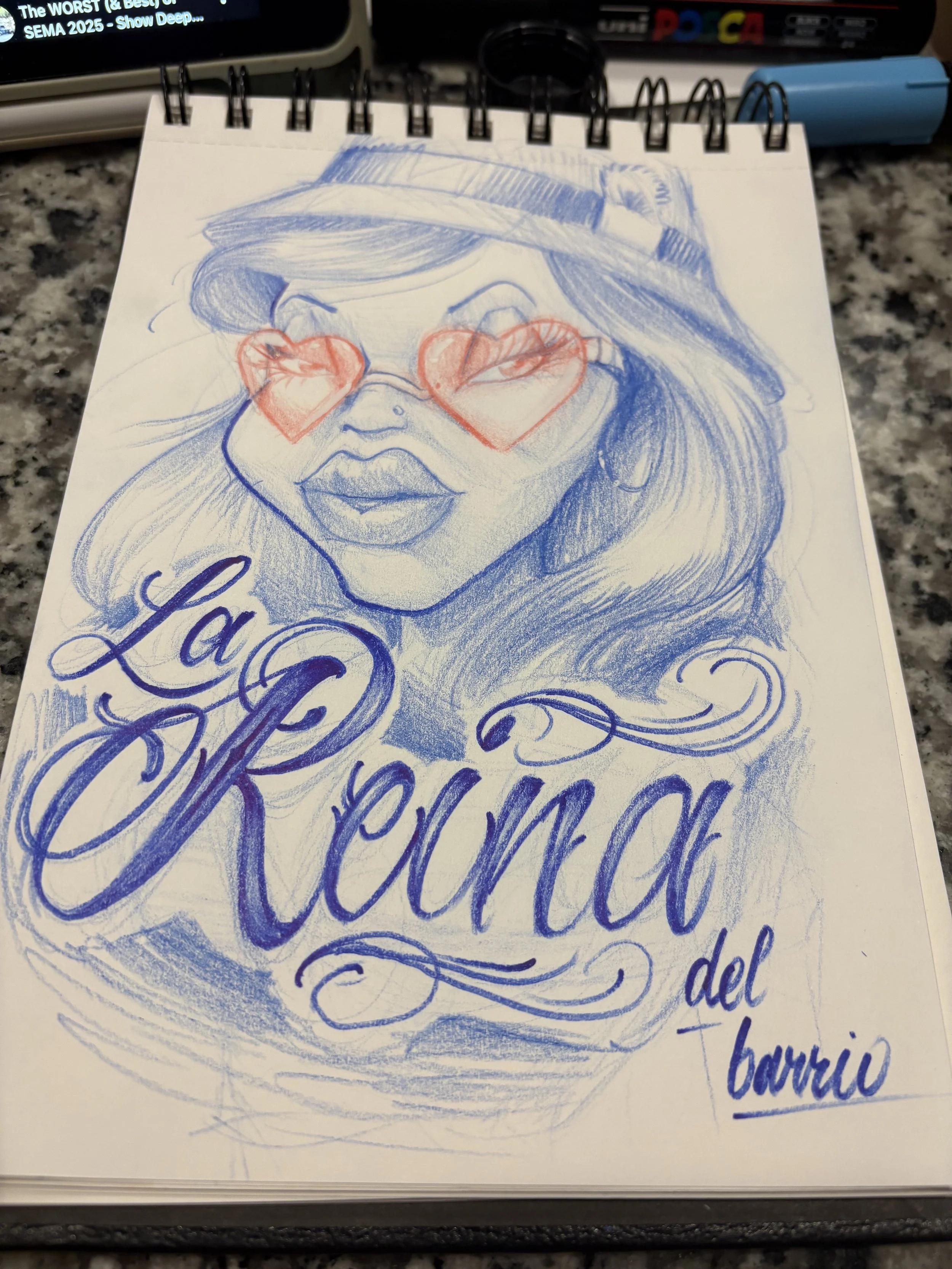 A sketch of a woman's face with long hair wearing a hat. She is holding her sunglasses, which are red hearts, in front of her eyes. The sketch is drawn in blue ink with some cursive writing below, and the background shows a computer monitor, markers, and other stationery on a speckled surface.