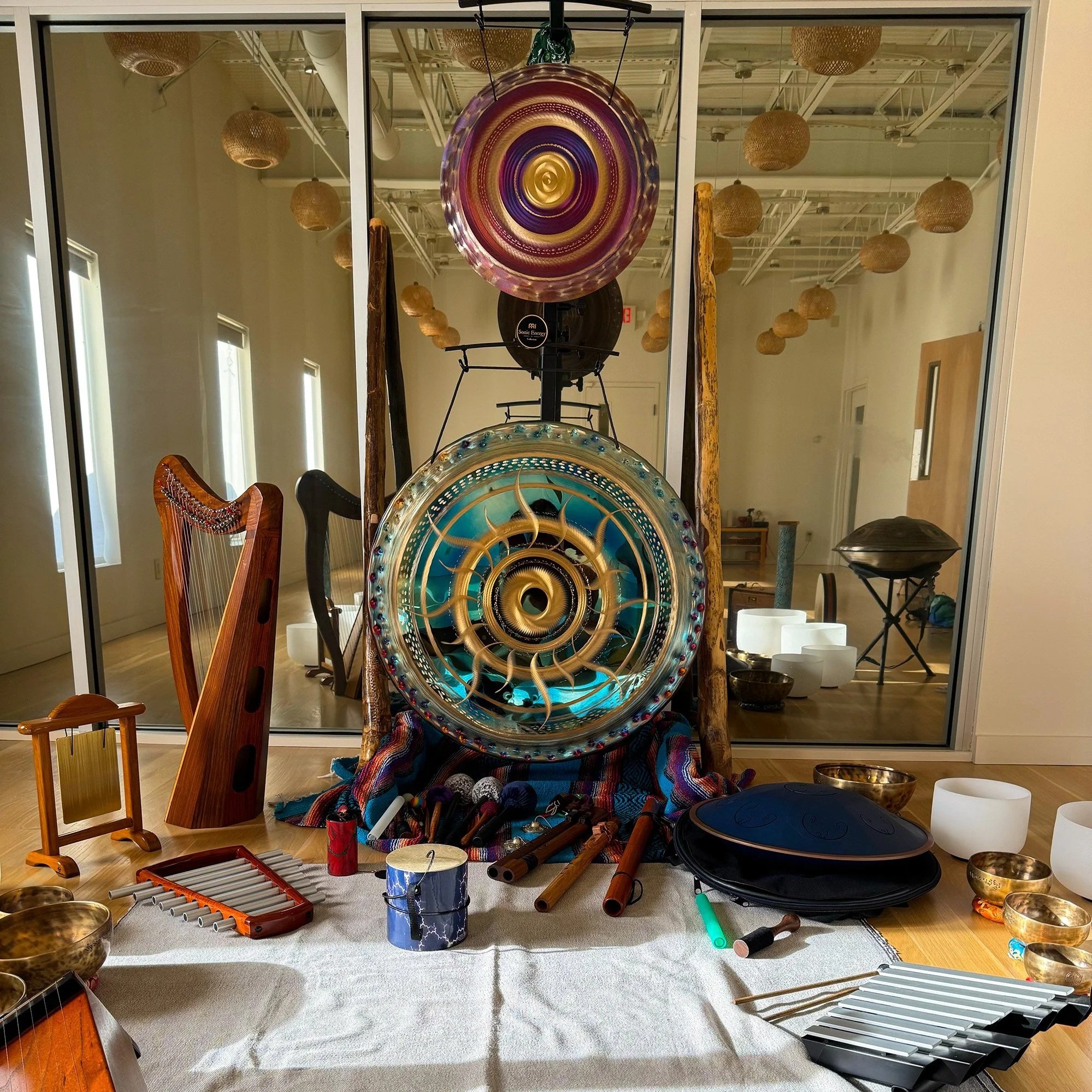Various musical instruments including a harp, wind chimes, and singing bowls arranged on a white cloth in front of a large glass display case with colorful hanging art pieces inside.