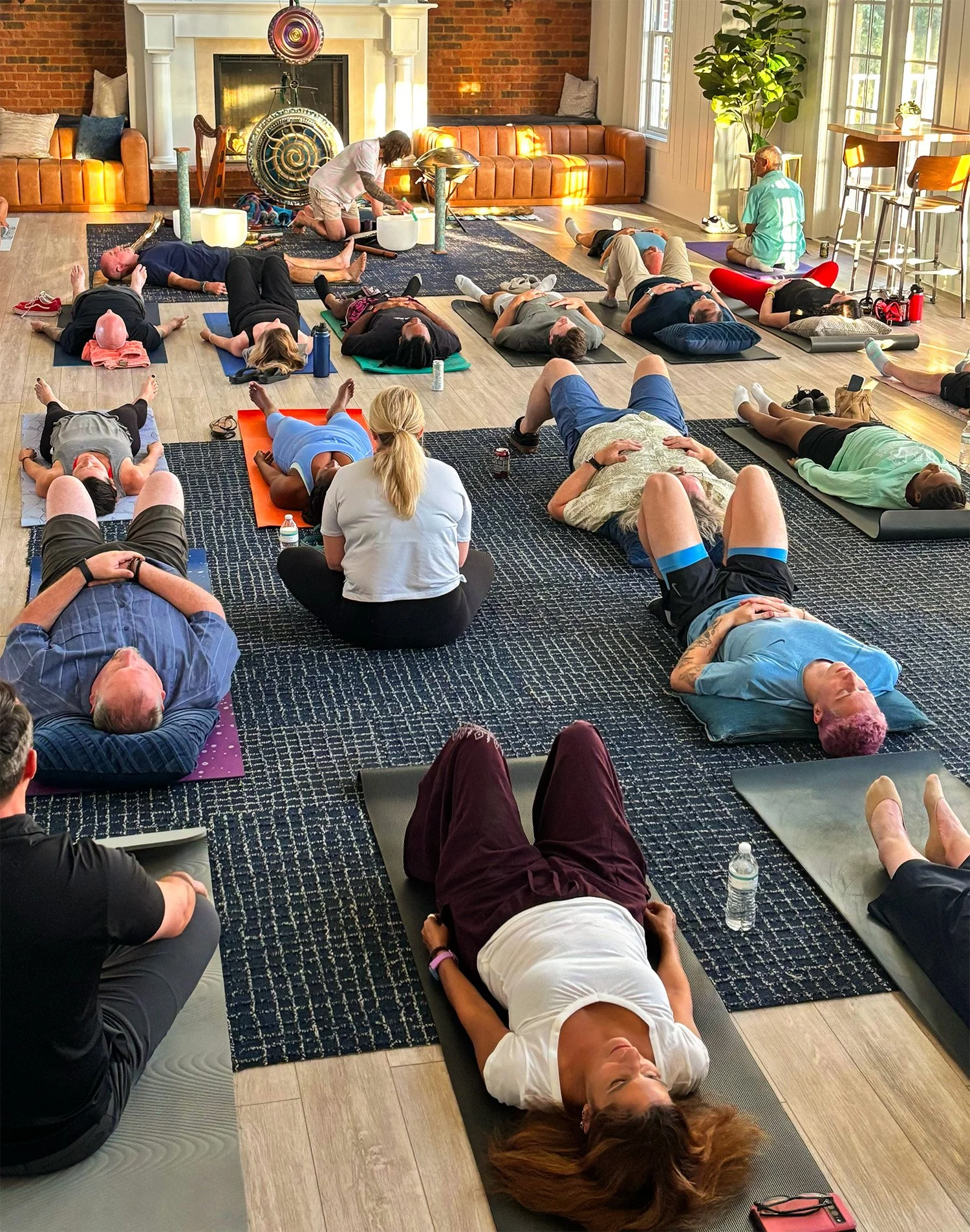People participating in a group yoga or meditation class in a bright, cozy living room with large windows, a fireplace, and a mix of colorful and neutral furniture.