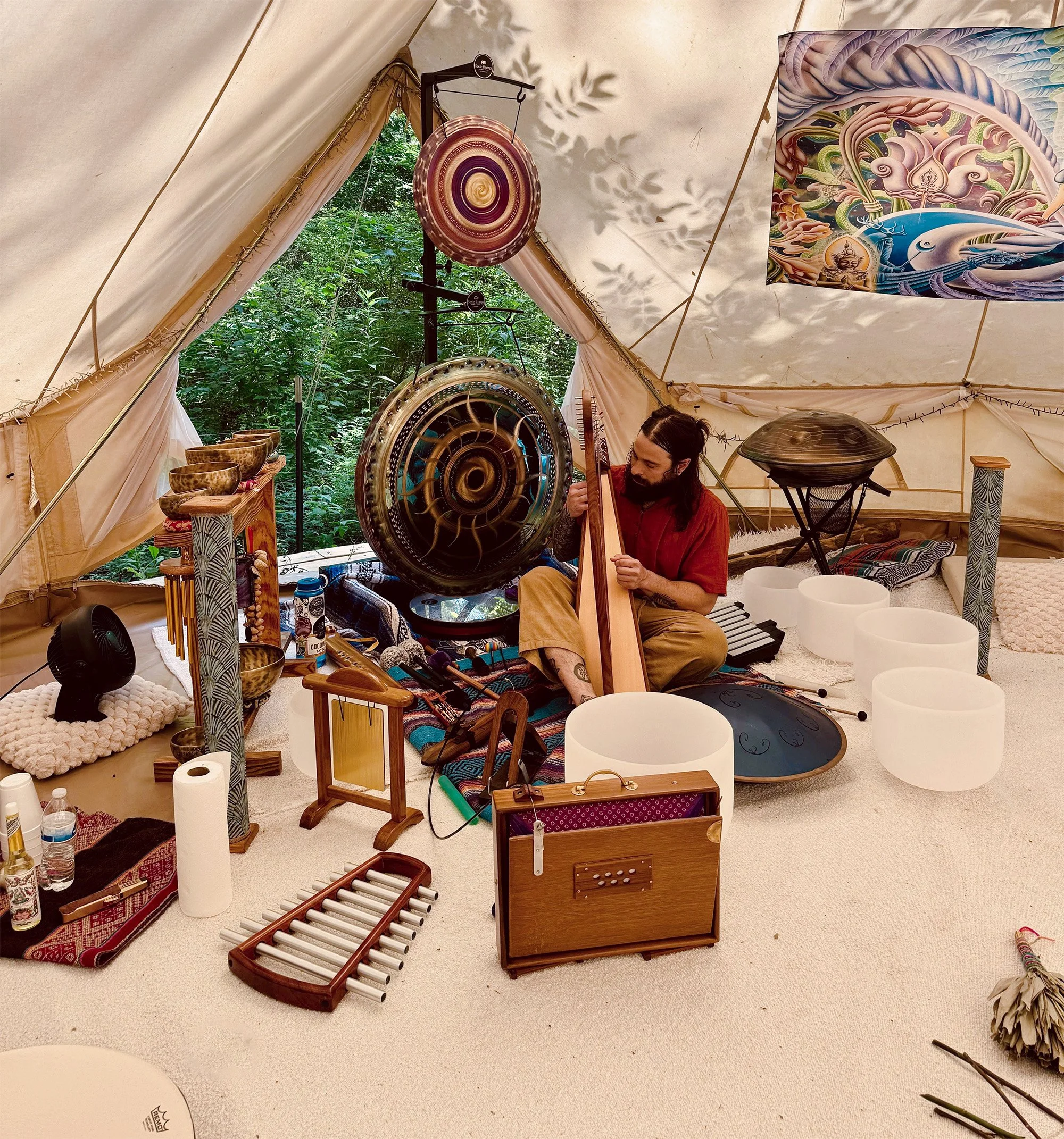 A man seated inside a beige tent filled with musical instruments, including gongs, chimes, and bowls, with a forest visible outside the open tent flap.