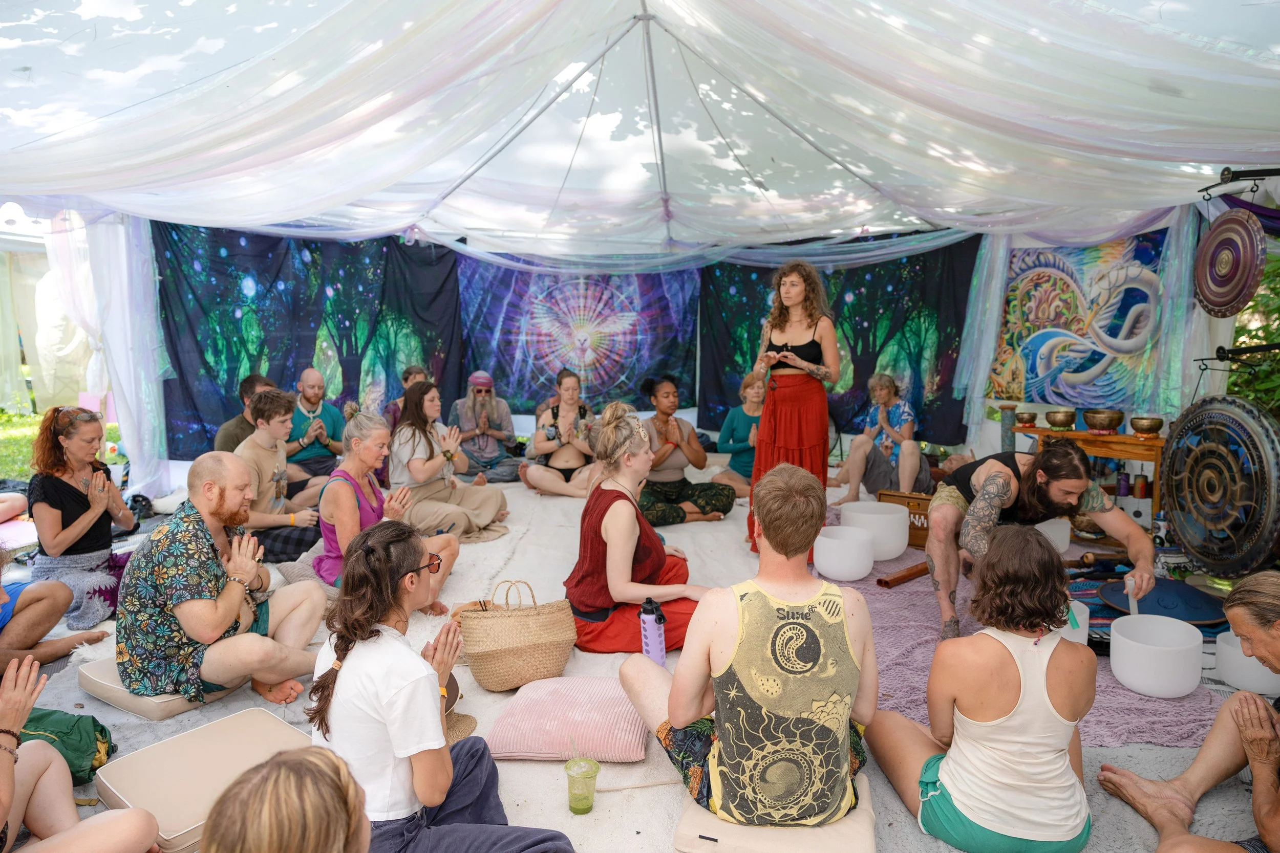 People participating in a meditation or sound healing session inside a decorated tent, sitting cross-legged with some holding hands in prayer, surrounded by musical instruments and vibrant wall hangings.