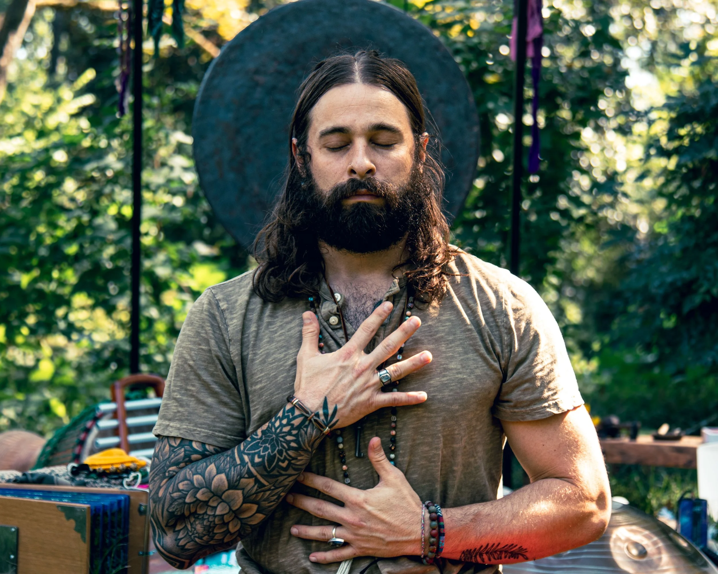 A man with long hair and a beard practicing meditation outdoors, standing with one hand on his chest and the other on his abdomen, with a serene expression and eyes closed, surrounded by lush green trees.