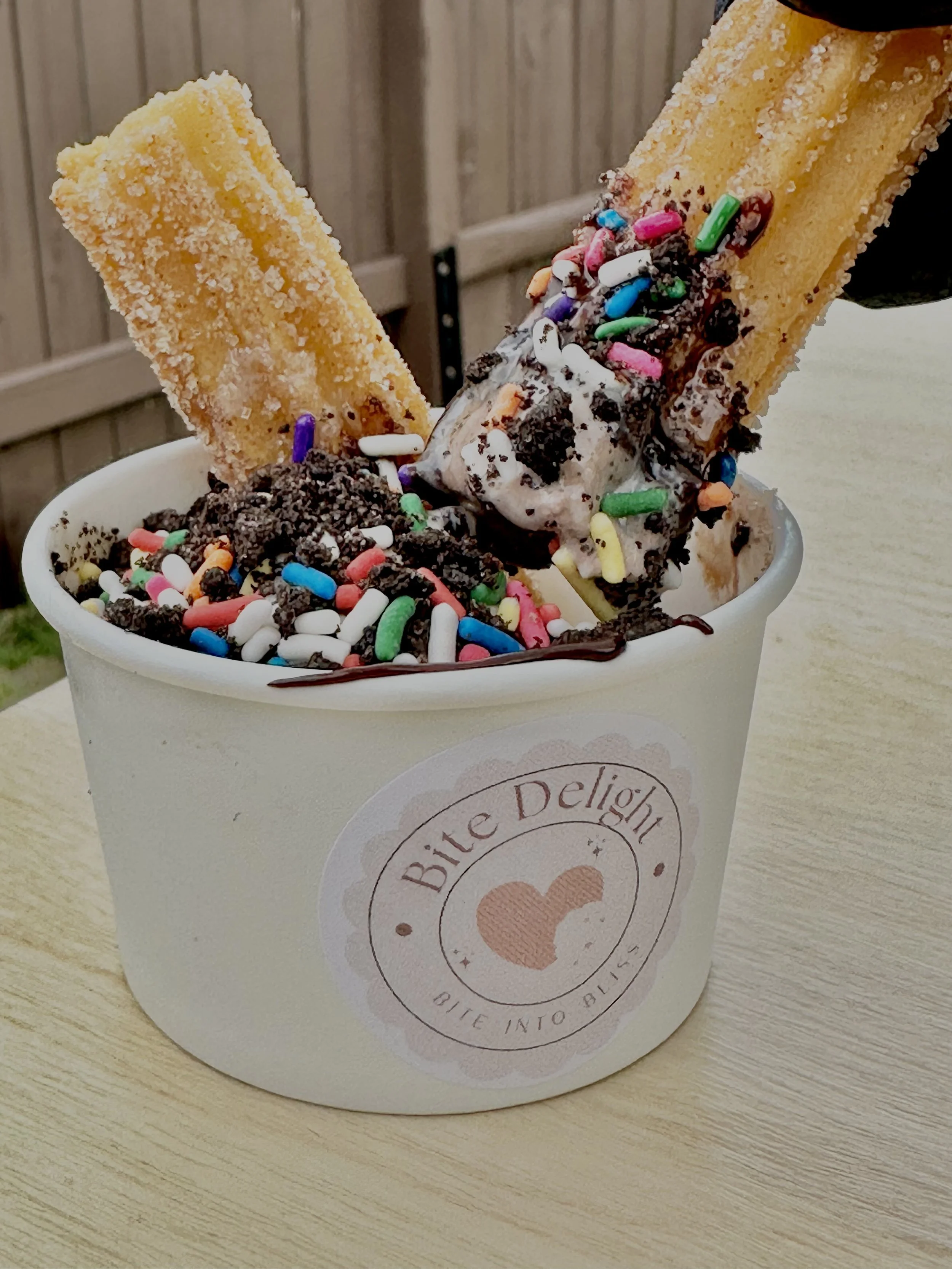 A cup of ice cream topped with sprinkles, cookies, and two biscuit sticks, with the cup labeled "Bite Delight".
