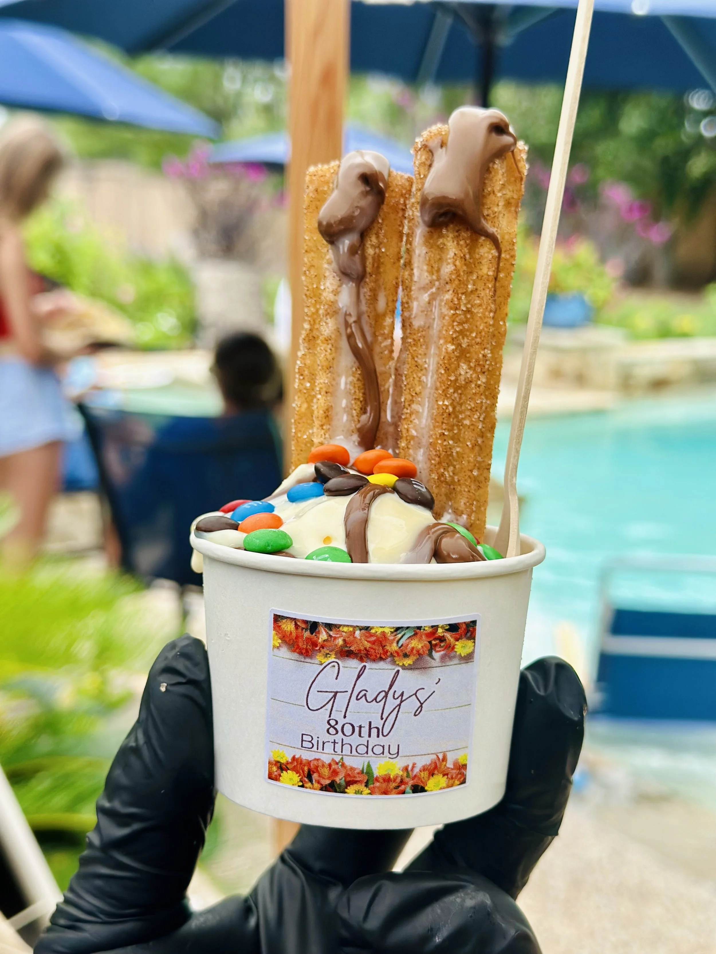 Cup of ice cream with colorful candies, two caramel cheesecake sticks with chocolate drizzle, and a label that reads 'Gladys' 80th Birthday', being held outdoors.