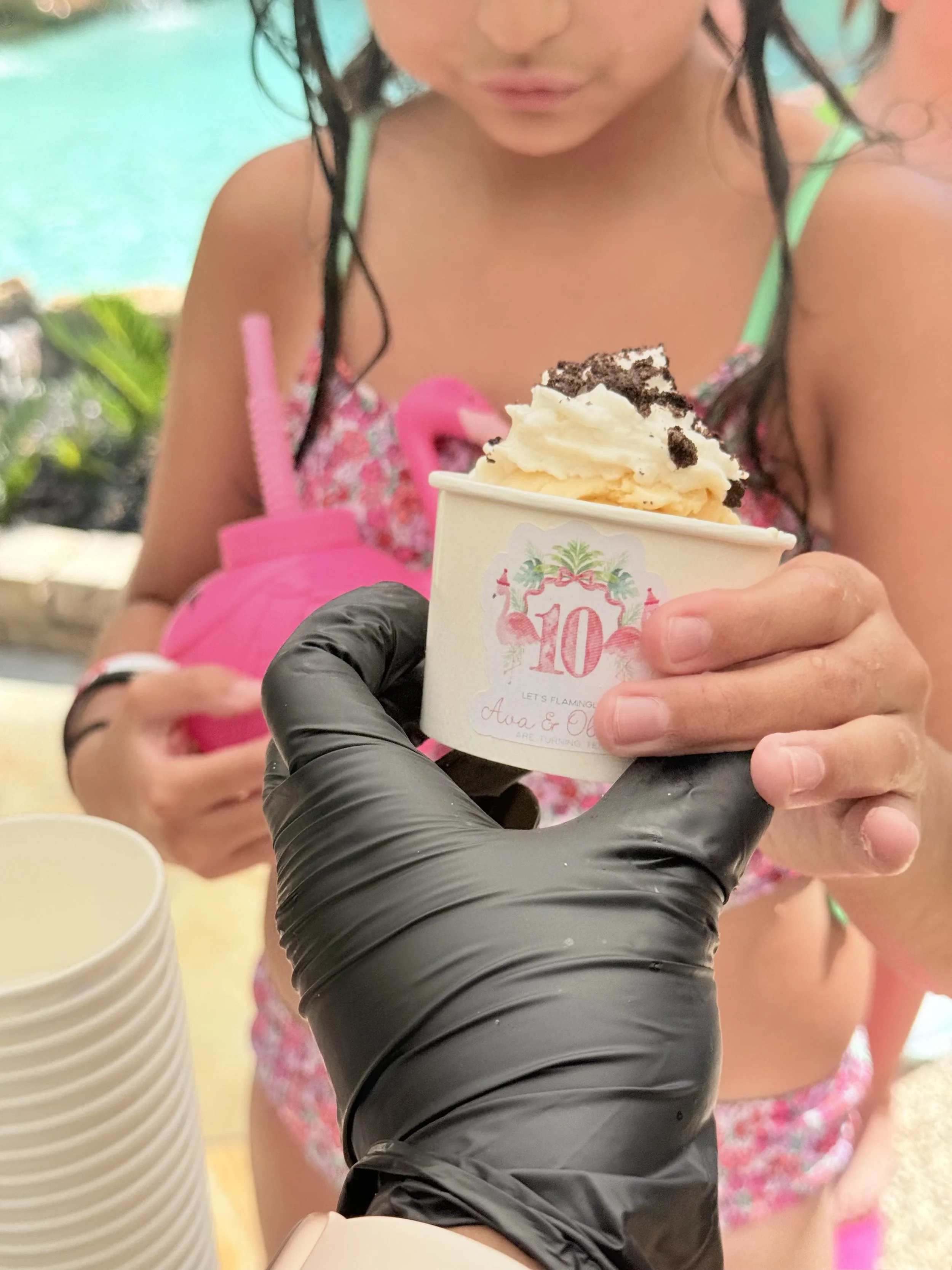 A girl in a swimsuit receives a cup of ice cream with whipped cream and cookie crumbs from a person wearing a black glove, at a poolside birthday celebration.