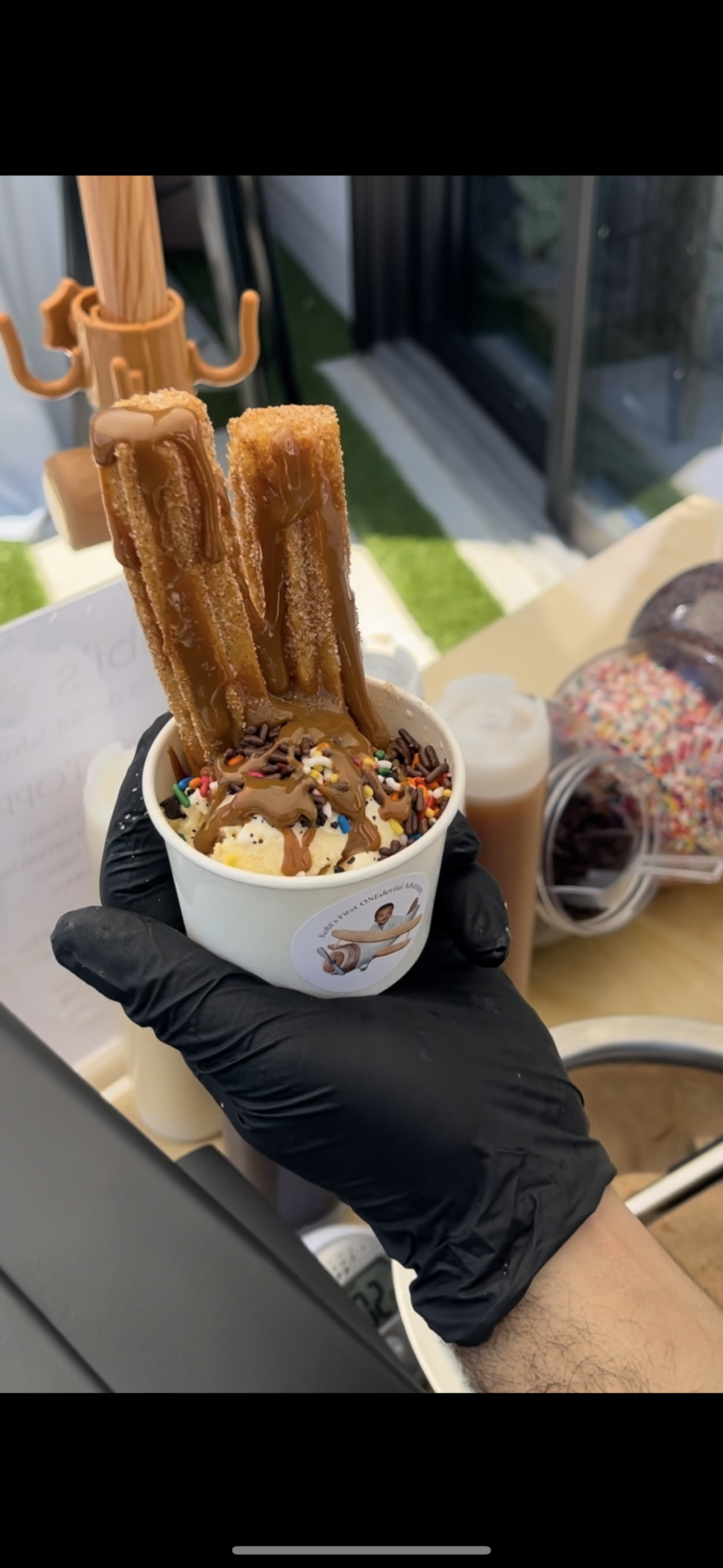 A person wearing a black glove holding a cup of ice cream topped with caramel sauce, chocolate sprinkles, and two crispy wafer sticks drizzled with caramel. In the background, there is a container of rainbow sprinkles and other toppings.