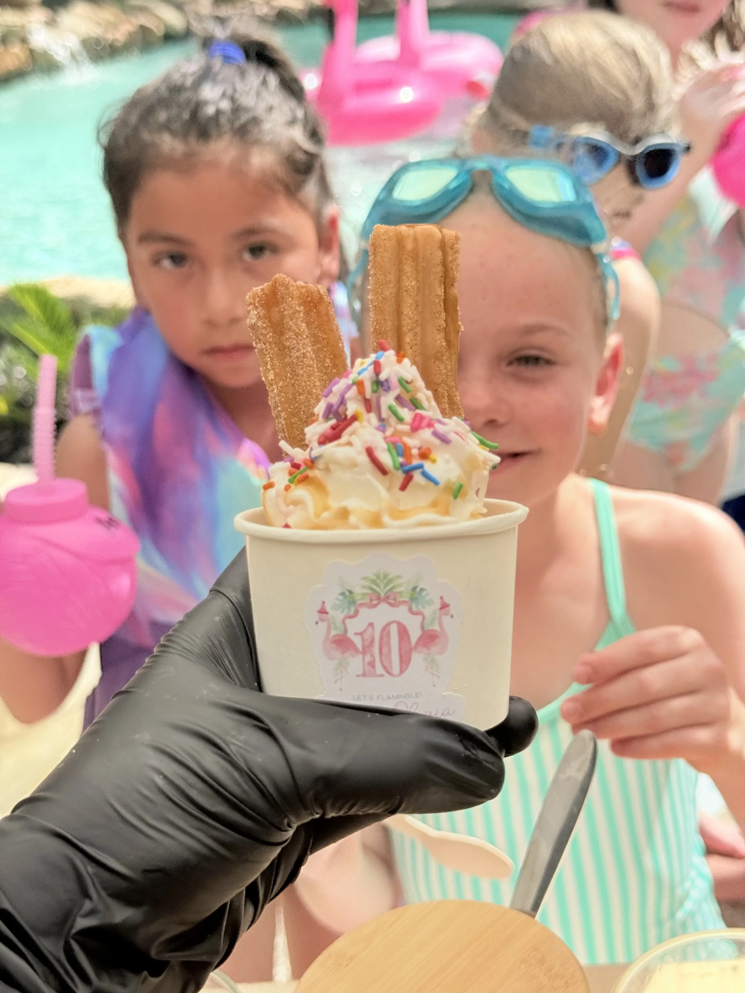 Children in swimsuits enjoying a pool, with one girl holding a cup of ice cream topped with sprinkles and cookies in front of the camera.